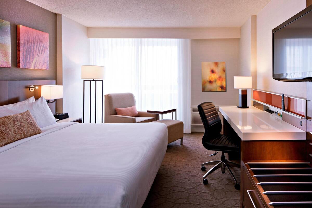 Quarto de hotel com uma cama grande, uma escrivaninha com cadeira, televisão e janela com cortinas.