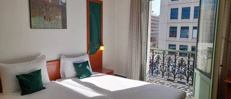 Ein Hotelzimmer mit einem großen Bett, einem Bild an der Wand, einem Fenster mit Blick auf Gebäude und einem Balkon mit schmiedeeisernem Geländer.