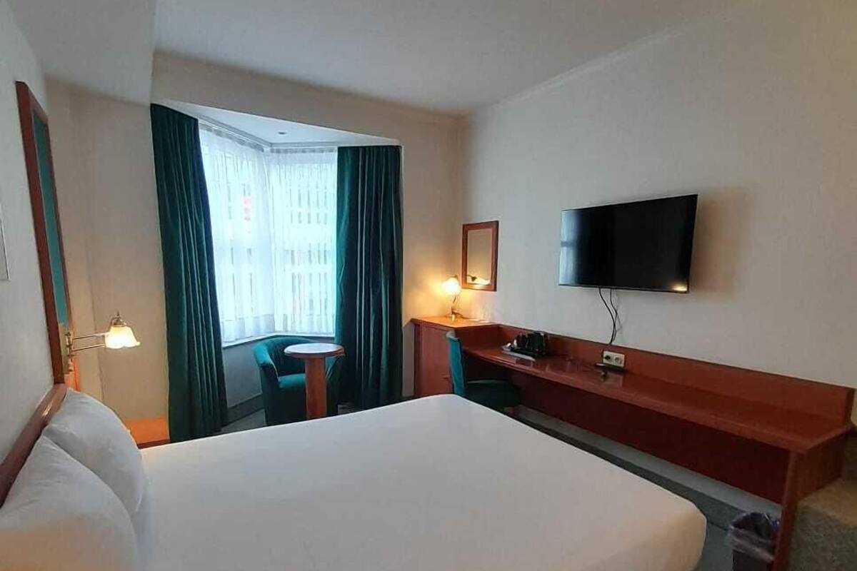 Habitación de hotel con una cama grande, un escritorio, una silla, un televisor y un ventanal con cortinas.
