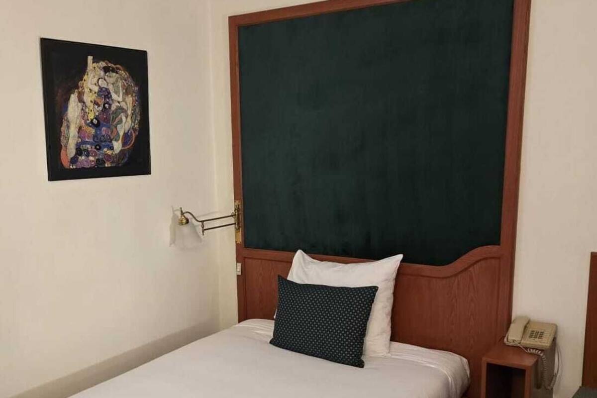Una habitación de hotel con una cama individual, un cabecero de madera, una mesita de noche con un teléfono y una pequeña mesa redonda.