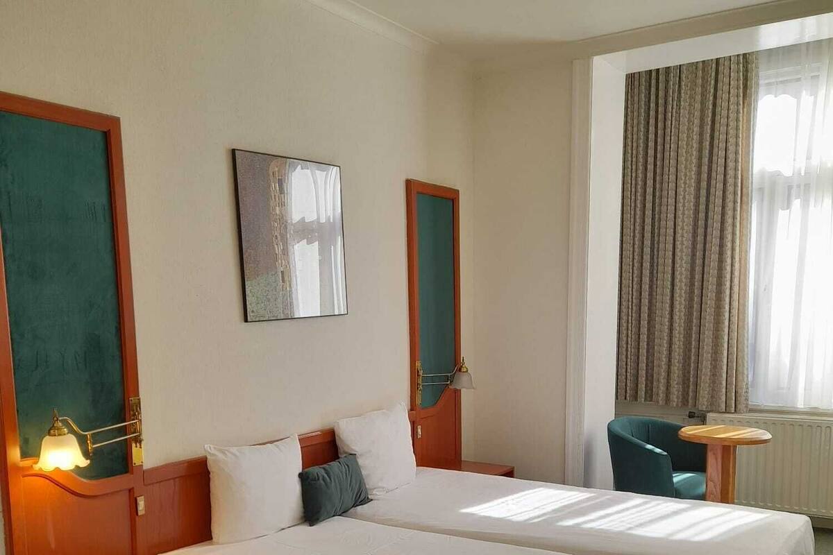 Habitación de hotel con dos camas, un escritorio, una silla, una lámpara y una ventana con cortinas.