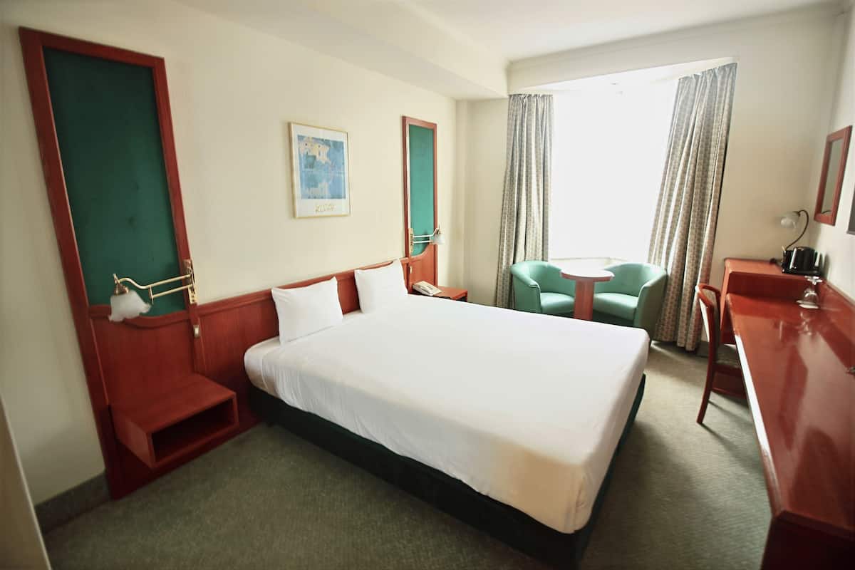 Habitación de hotel con una cama grande, un escritorio, una silla y una mesita. Se ve una ventana con cortinas y un cuadro en la pared.