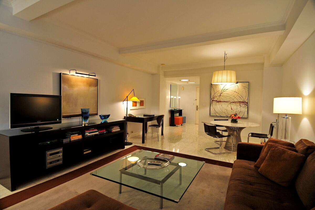 A modern living room with a flat-screen TV, a glass coffee table, a brown sofa, and a dining area with a table and chairs.