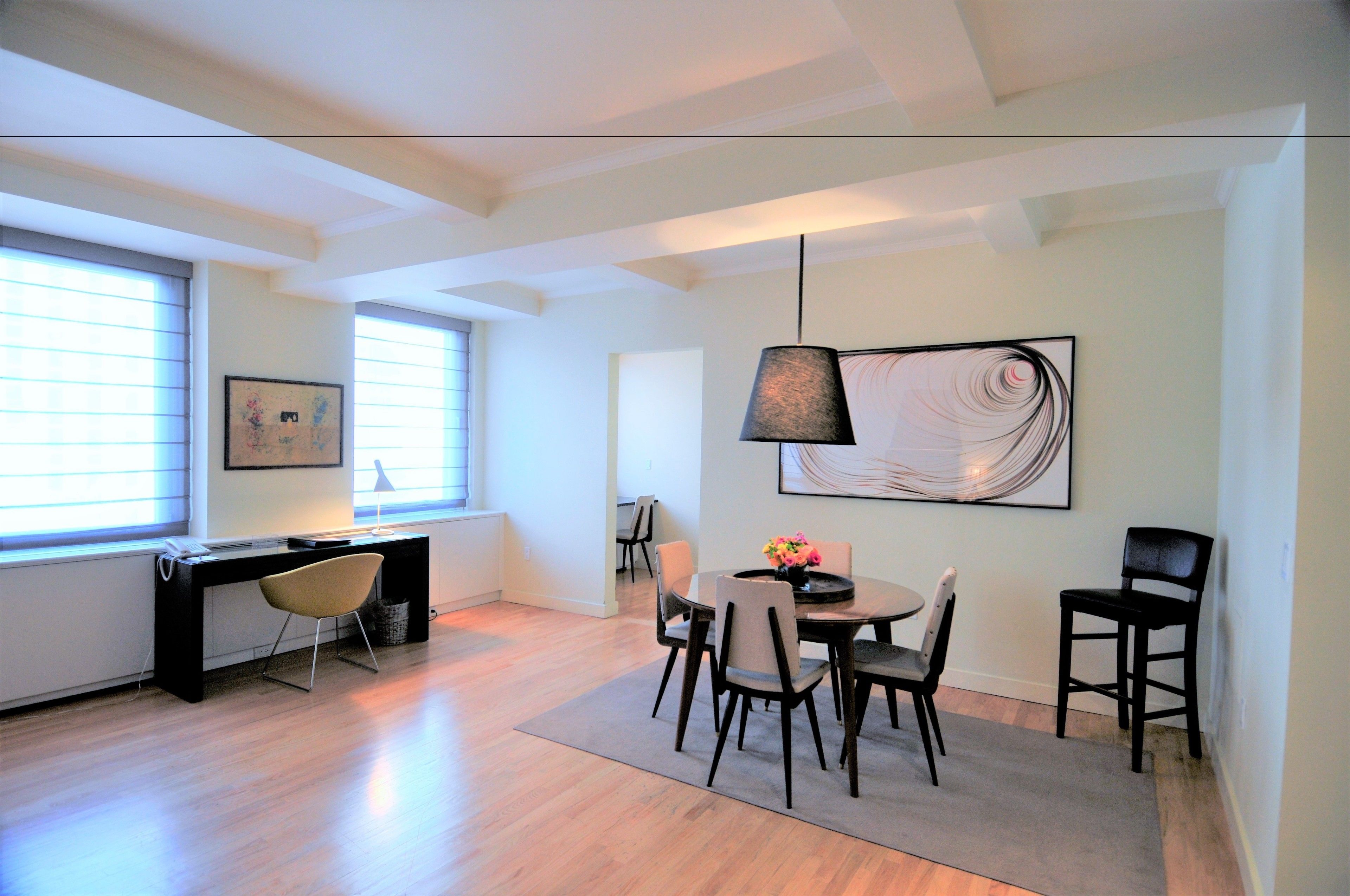 A modern living room with a dining area, featuring a black desk, a round dining table with chairs, and a large abstract painting on the wall.