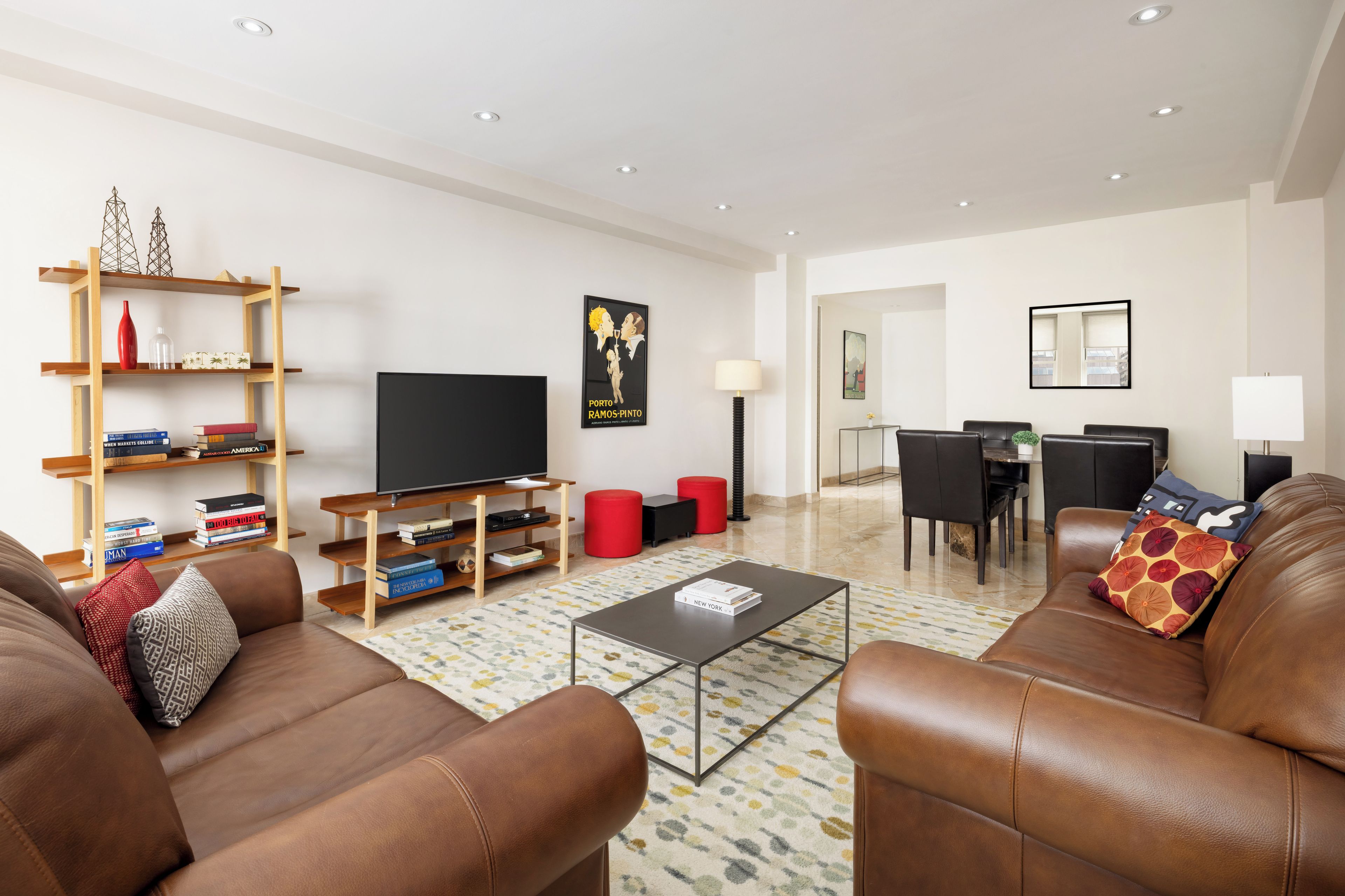 A modern living room with leather sofas, a flat-screen TV, wooden shelving with books, and a dining area with a table and chairs.