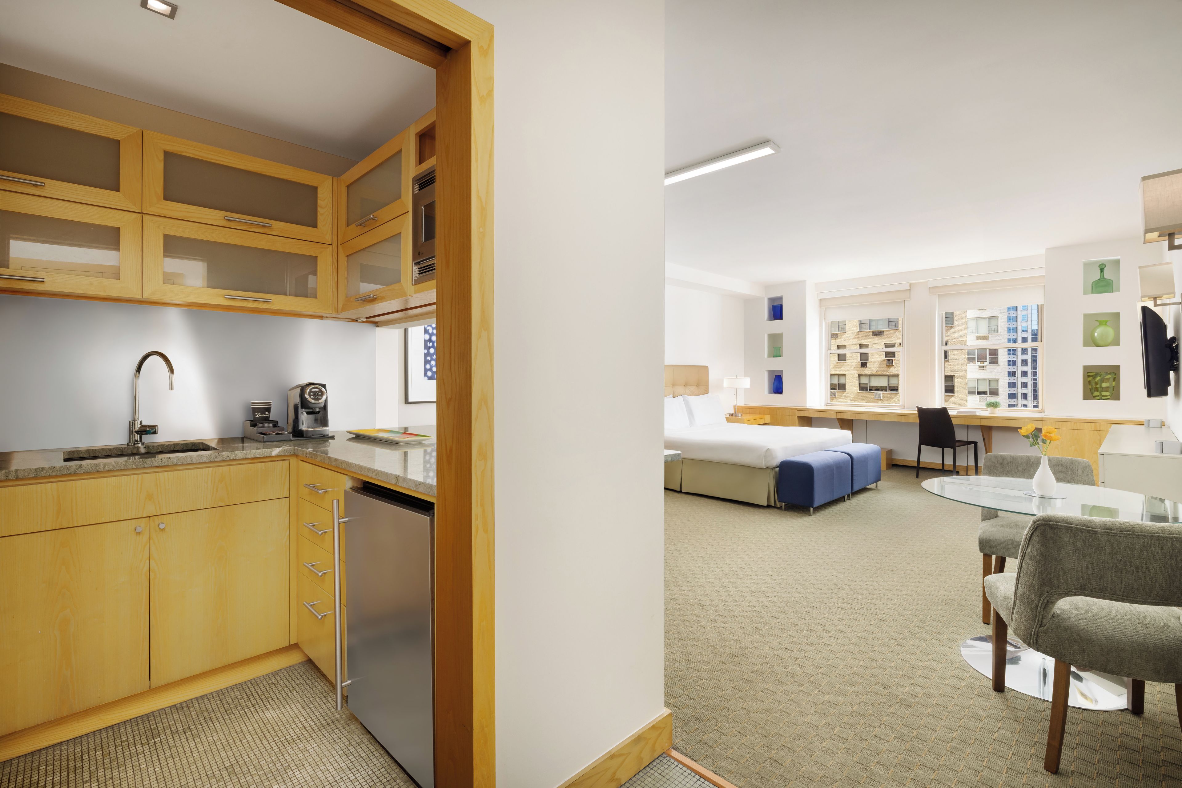 Een moderne hotelkamer met kitchenette, een bed met blauwe accenten, een bureau met stoel, en uitzicht op de stad.