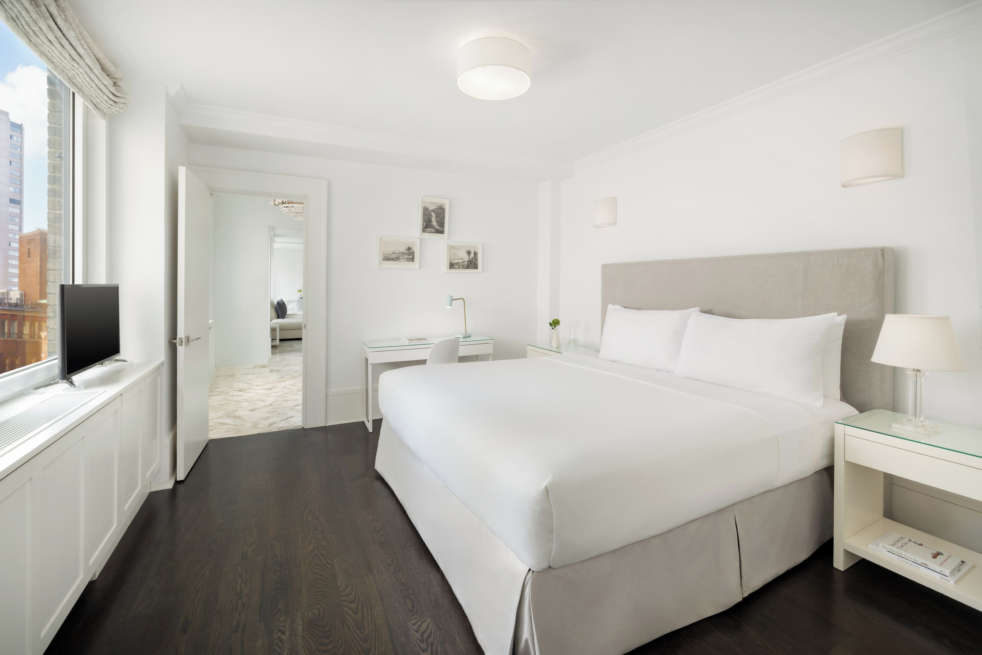 A hotel room with a large bed, a desk, a chair, a TV, and a window with a view of buildings.