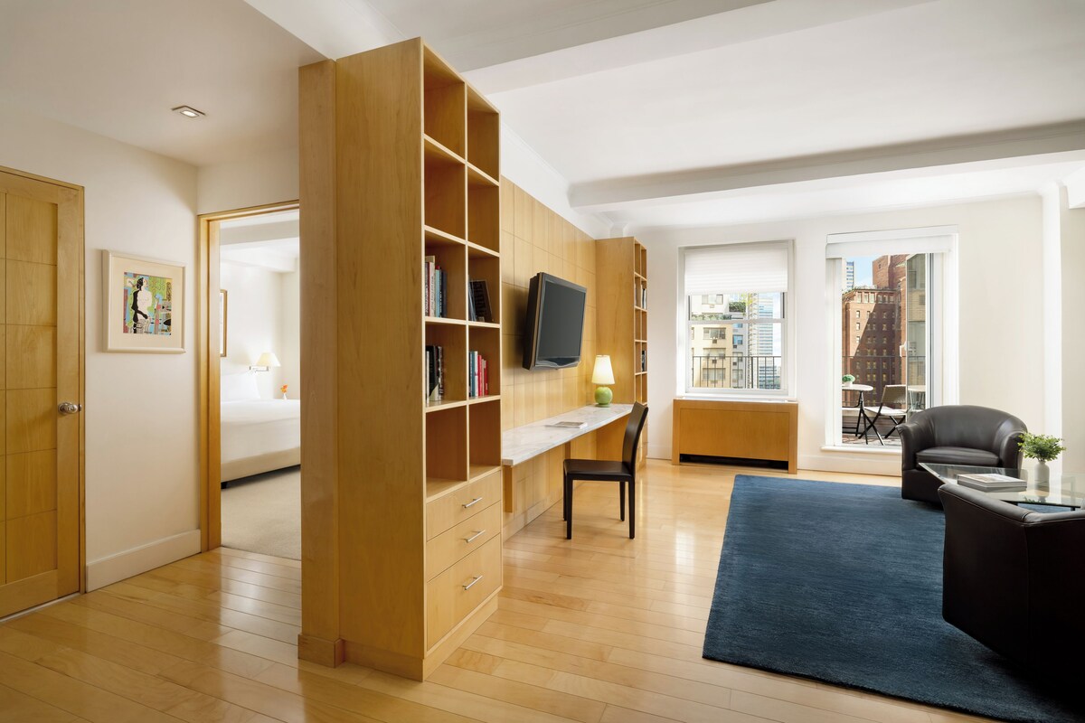 A modern living room with a wooden bookshelf, a flat-screen TV, a dining area with a table and chairs, and a view of the city through large windows.