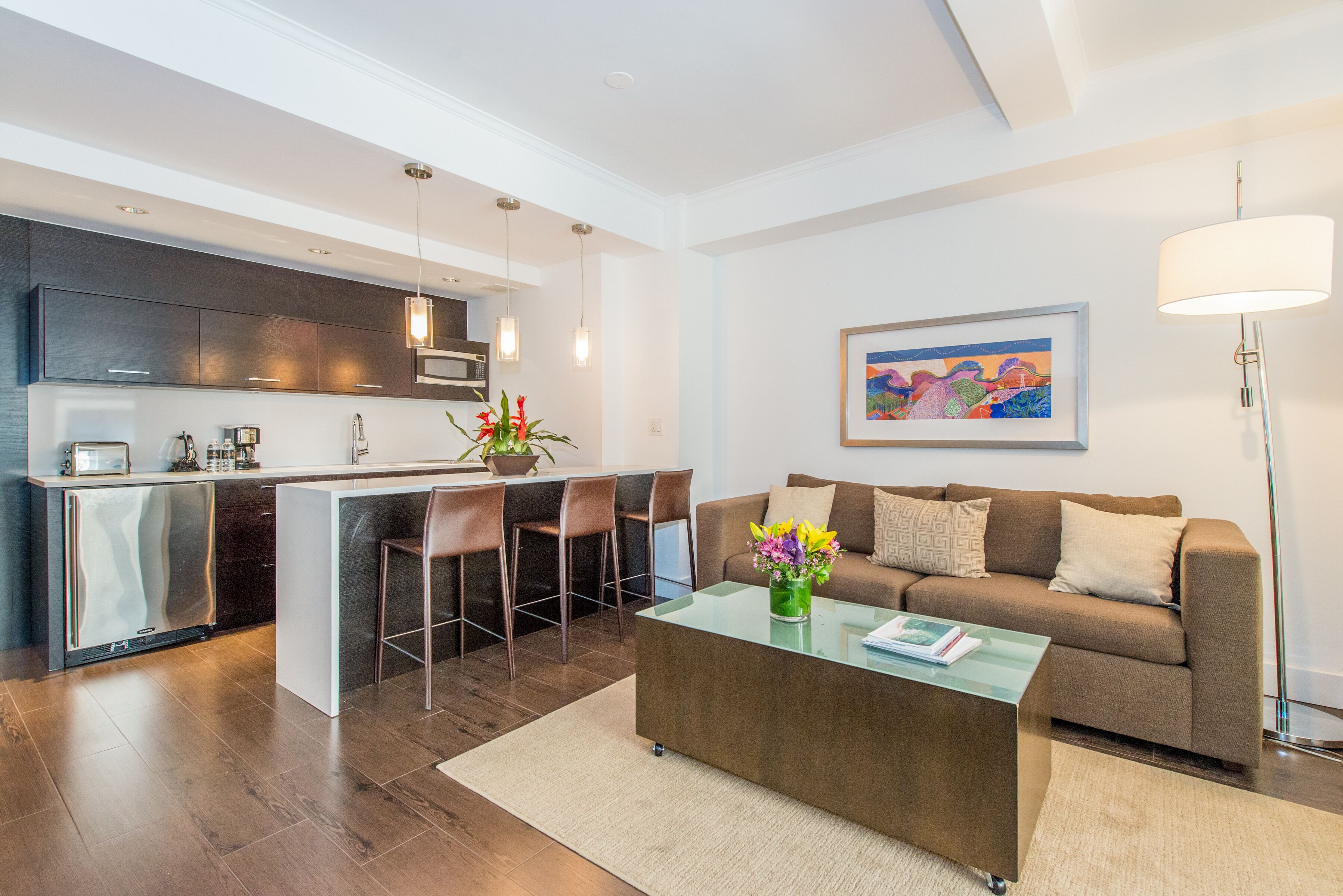 A modern kitchen with a central island, a dining area with bar stools, a sofa, and a coffee table with a vase of flowers.