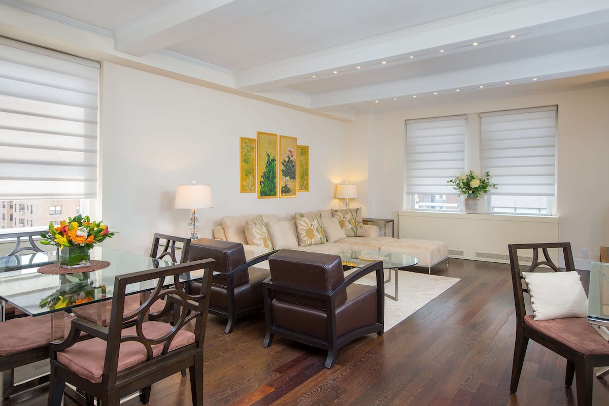 A modern living room with a glass dining table, dark brown chairs, a beige sofa, and a large window with blinds.