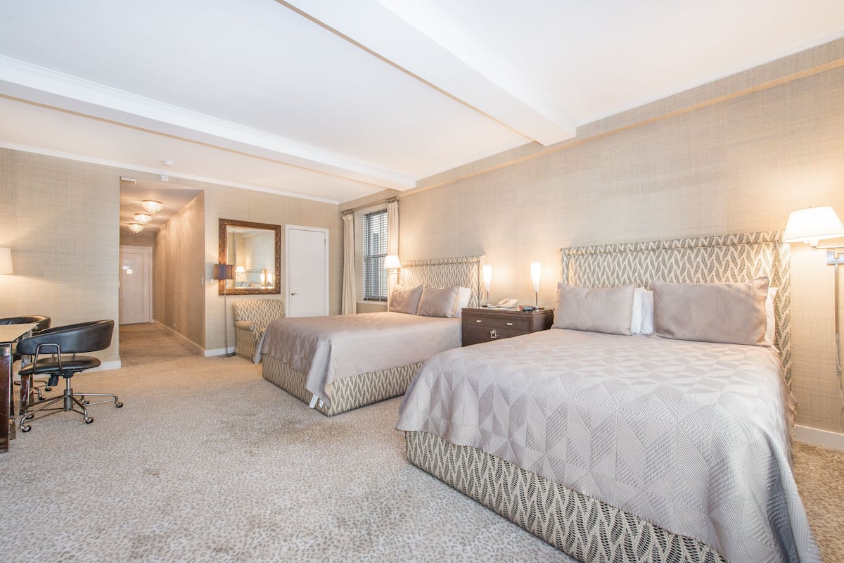A hotel room with two beds, a desk, and a chair. There is a door leading to another room and a window with curtains.