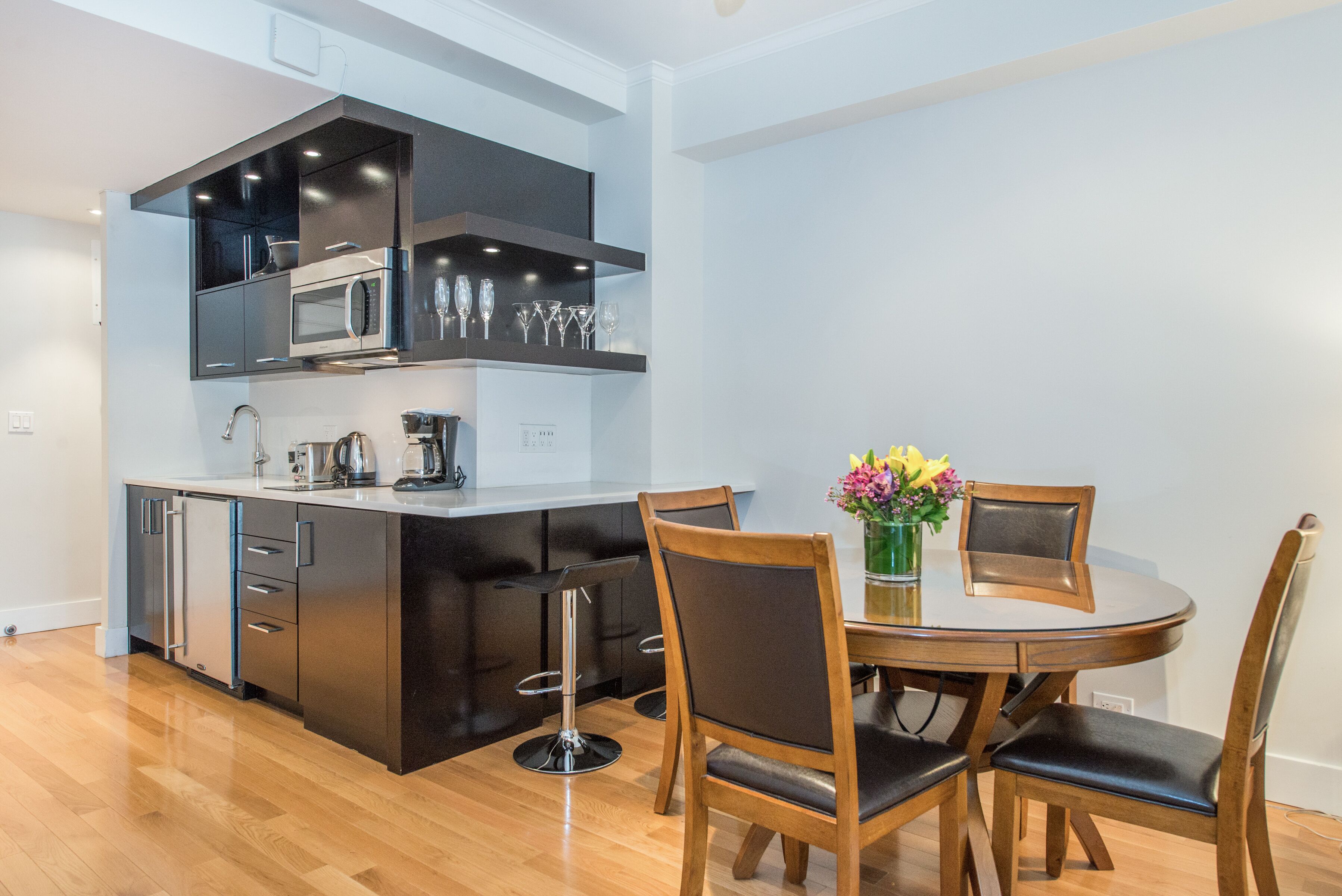 A modern kitchen with a built-in microwave, oven, and coffee maker. There is a dining table with chairs and a vase of flowers.