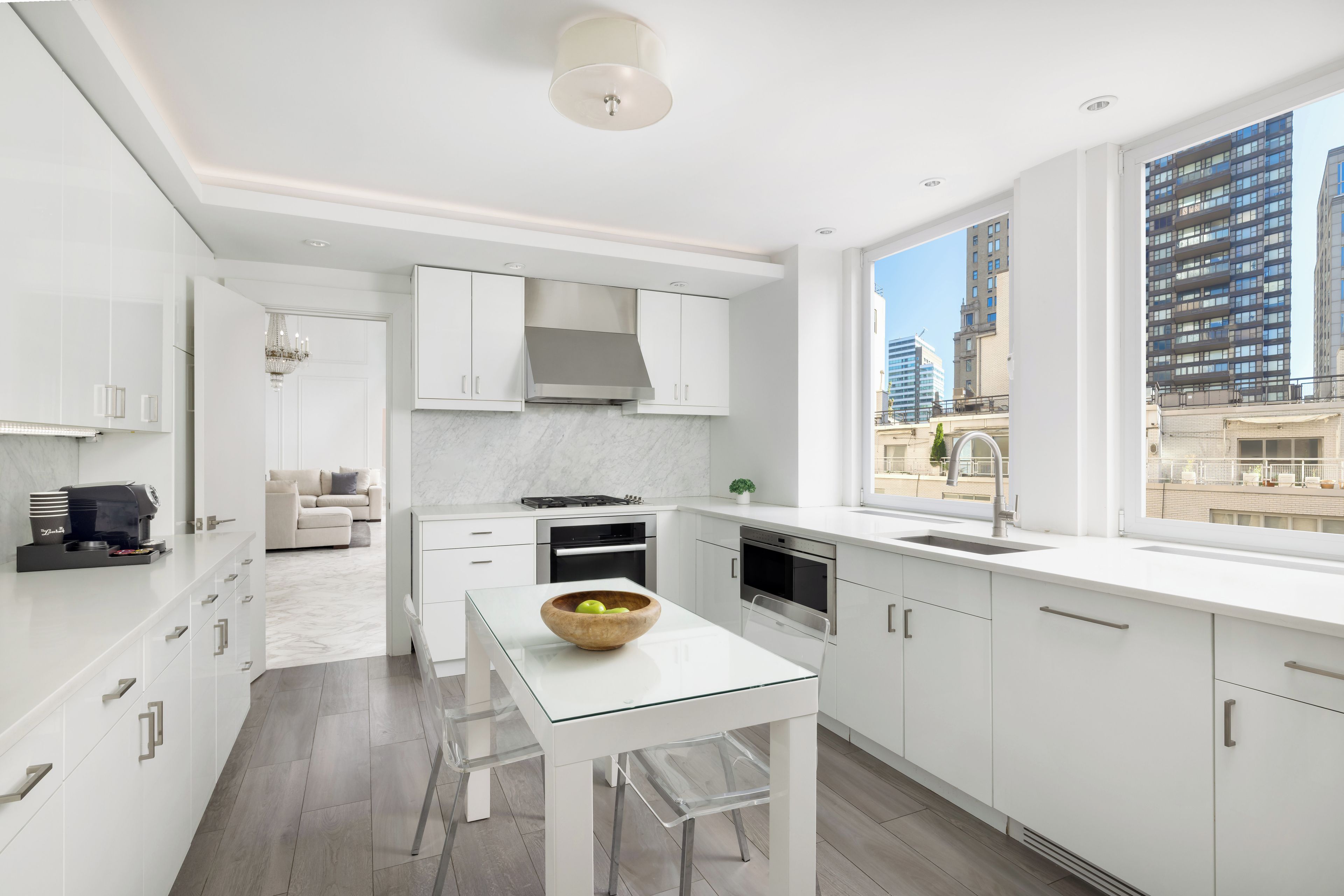 Una cocina moderna con armarios blancos, una mesa de comedor de vidrio y vistas a un paisaje urbano a través de un amplio ventanal.