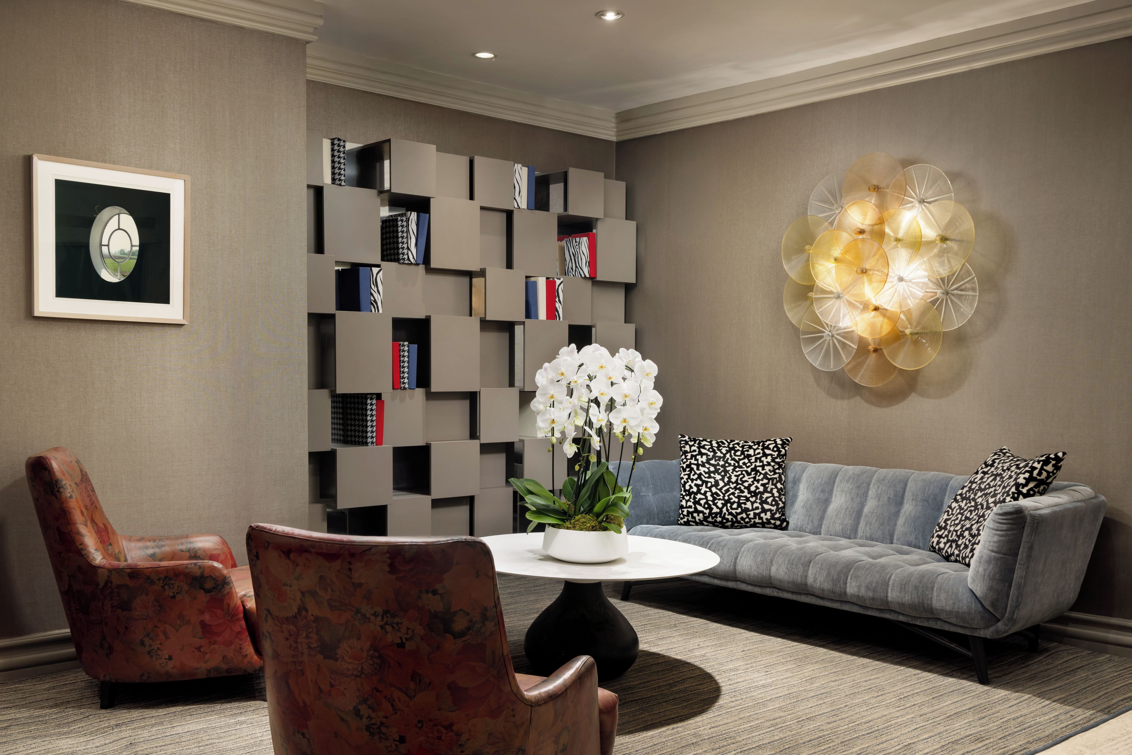 A modern living room with a grey sofa, floral armchairs, a round coffee table, a bookshelf, and a decorative wall piece.