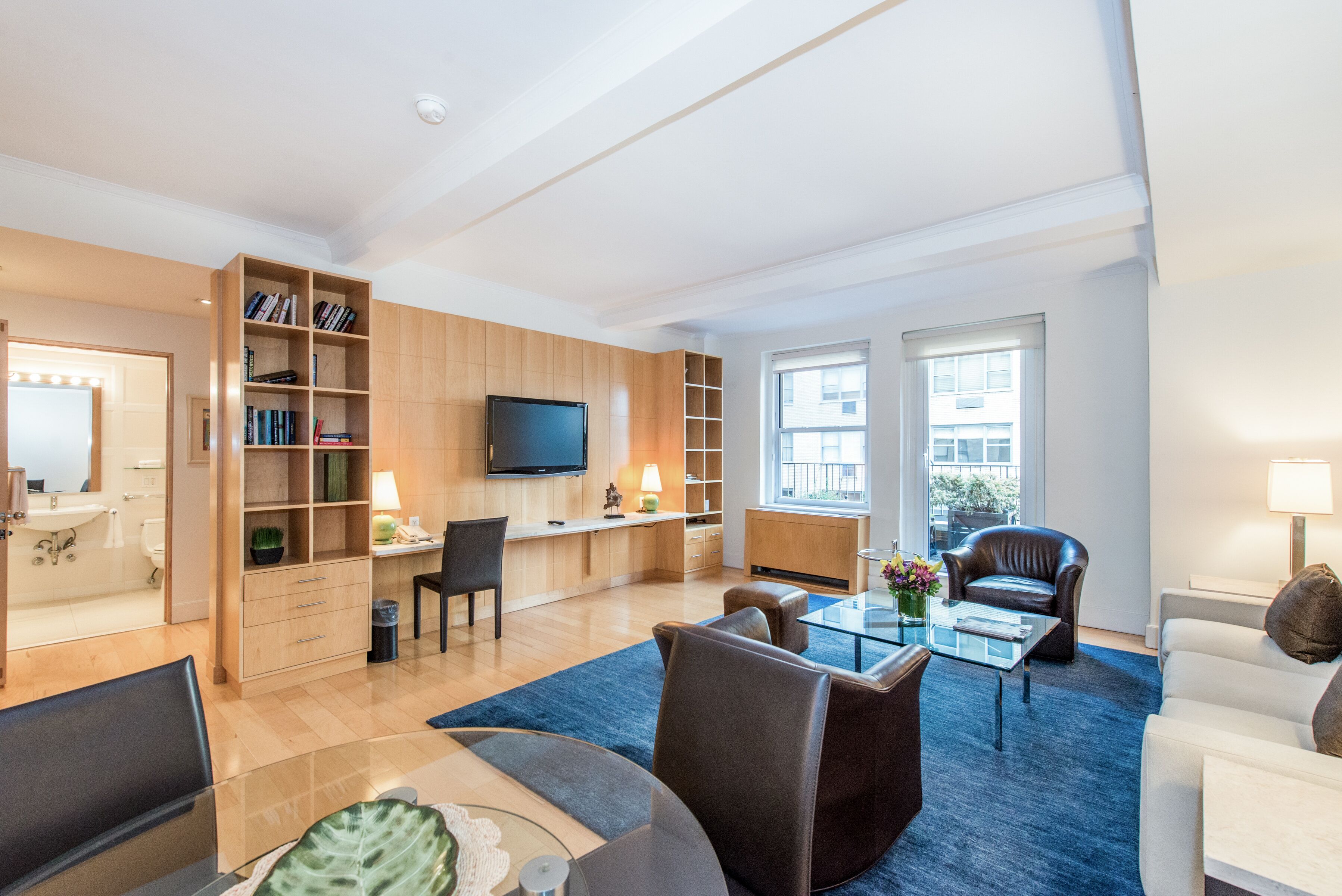 Een moderne woonkamer met een flatscreen televisie, een glazen salontafel en een bank. Er is een boekenkast met boeken en een lamp.