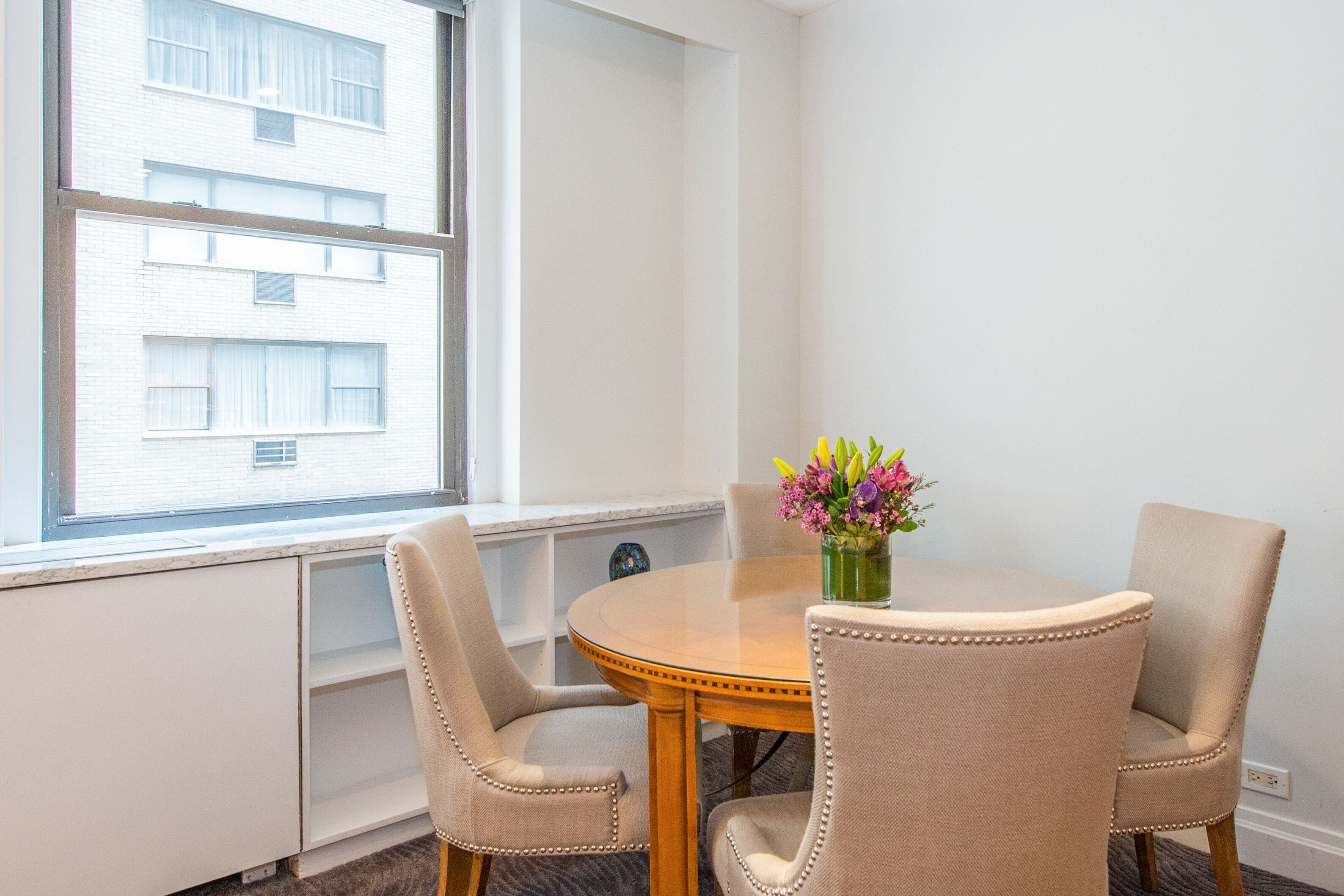 Comedor con una mesa redonda, cuatro sillas color beige con tapicería y un jarrón con flores.