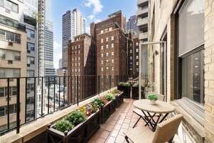 A balcony with a long planter filled with flowers, a wicker table, and chairs.