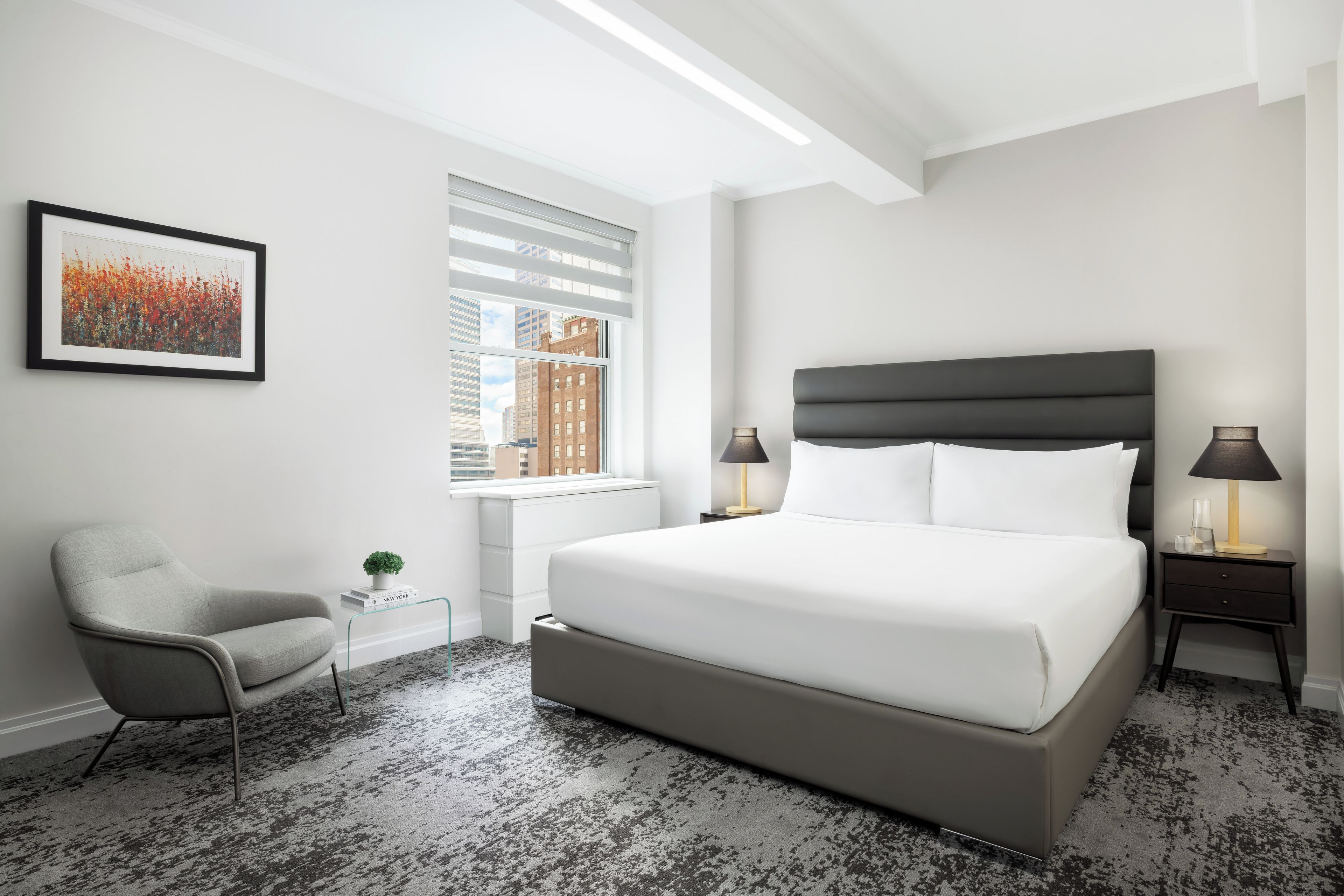 A modern hotel room with a large bed, a chair, a bedside table with a lamp, and a window with a city view.