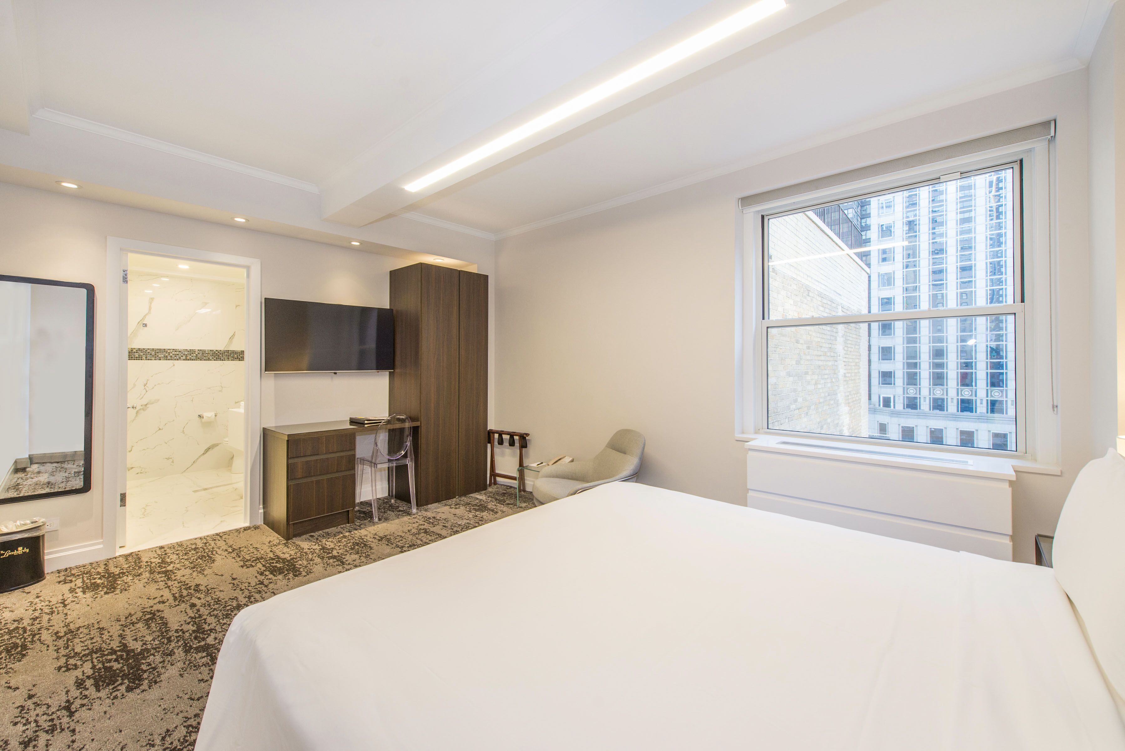 Ein modernes Hotelzimmer mit einem großen Bett, einem an der Wand befestigten Fernseher, einer Sitzecke mit einem Sessel und einem Fenster mit Blick auf die Stadt.