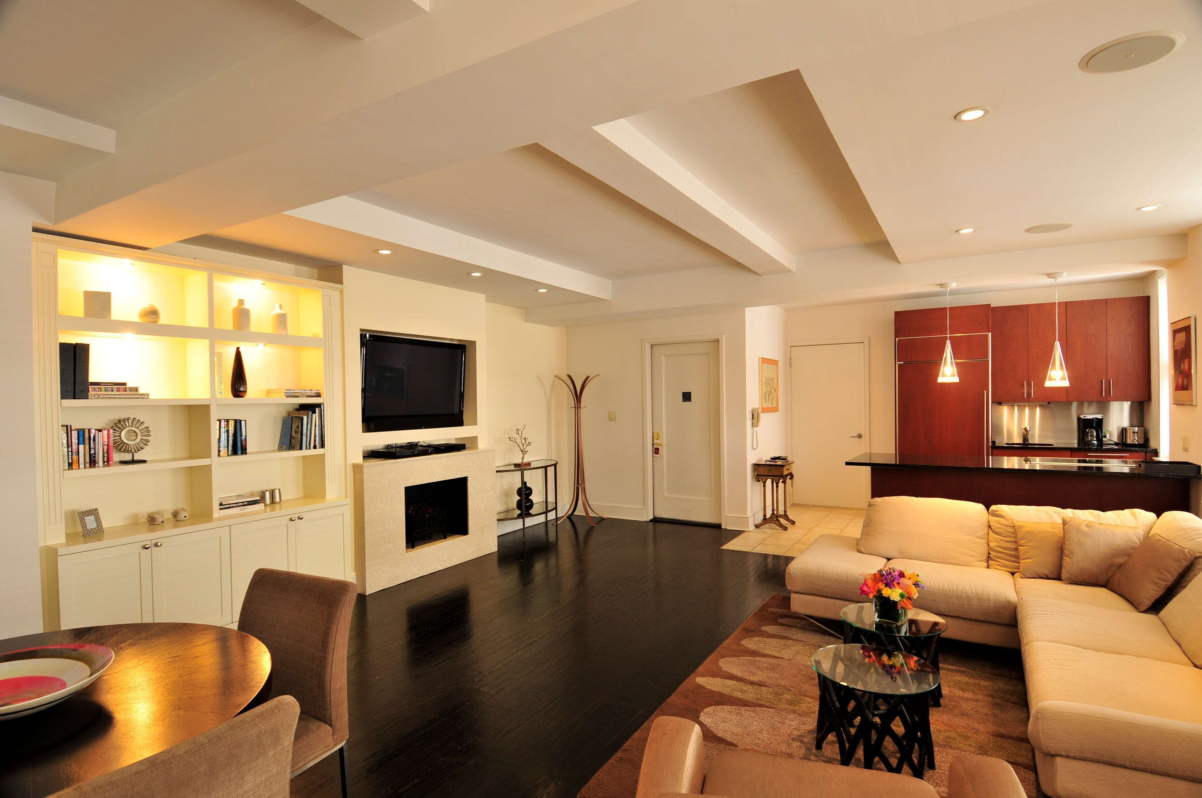 A modern living room with a sofa, a coffee table, and a TV mounted on the wall. There is a kitchen area with cabinets and a countertop visible in the background.