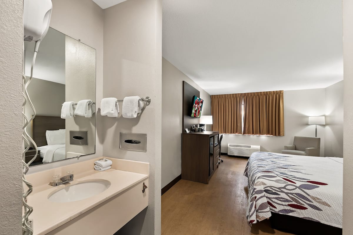 Ein Hotelzimmer mit großem Spiegel, Waschbecken, einem Bett mit Blumenmuster auf der Bettdecke, einem Schminktisch, einem Schreibtisch mit Lampe und einem Fenster mit Vorhängen.