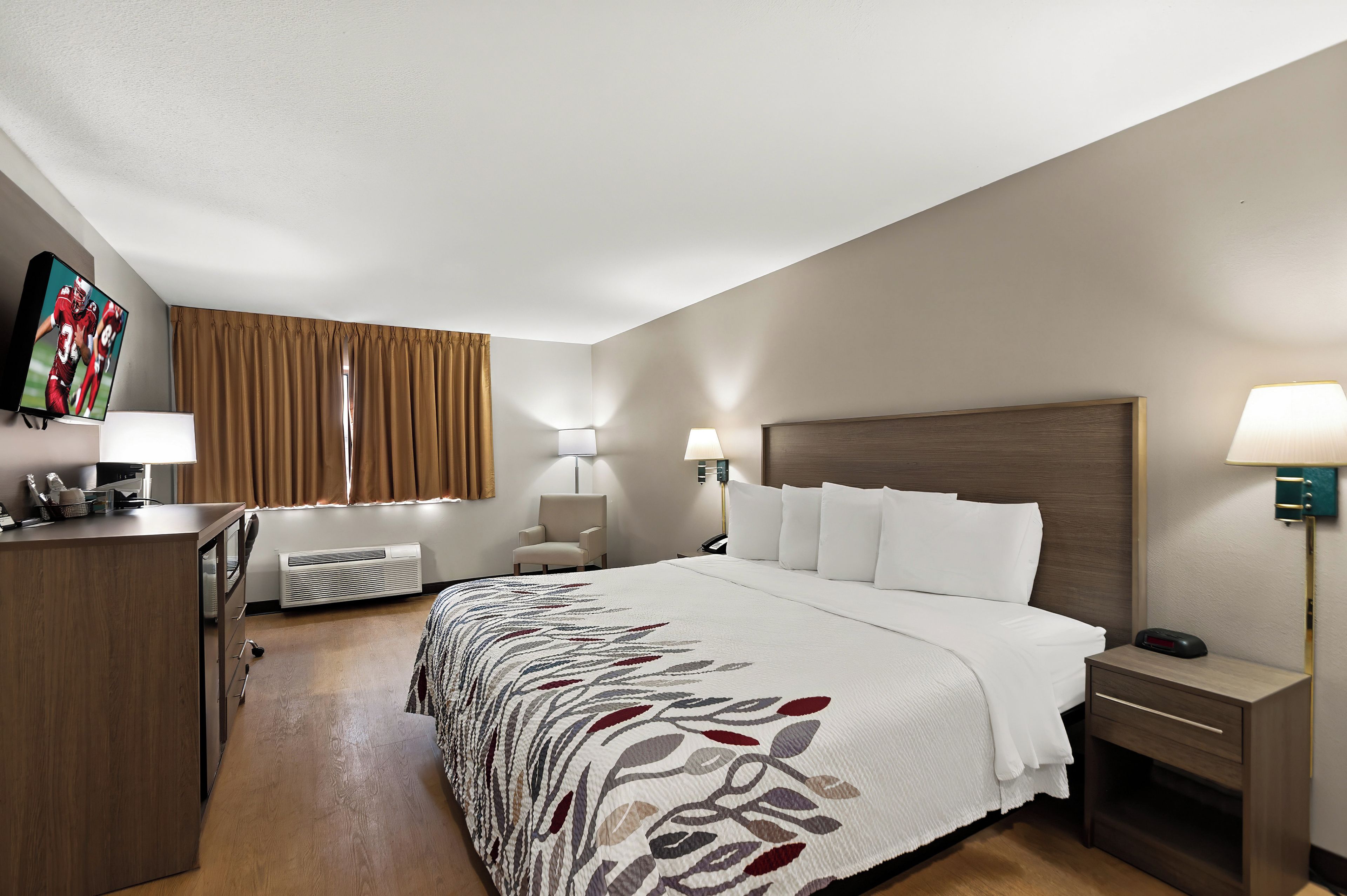 Quarto de hotel com cama, abajures, escrivaninha, cadeira e televisão na parede.