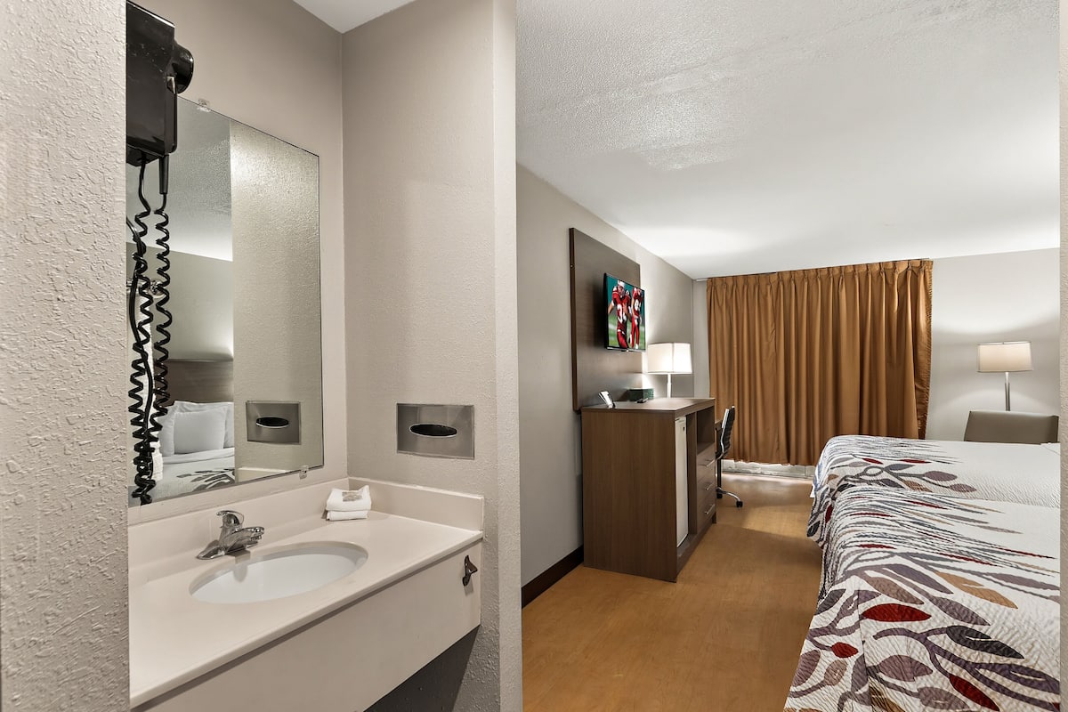 Ein Hotelzimmer mit Bad, das ein Waschbecken, einen Spiegel und einen Handtuchhalter umfasst. Das Zimmer verfügt über ein Bett, einen Schreibtisch und einen Stuhl.