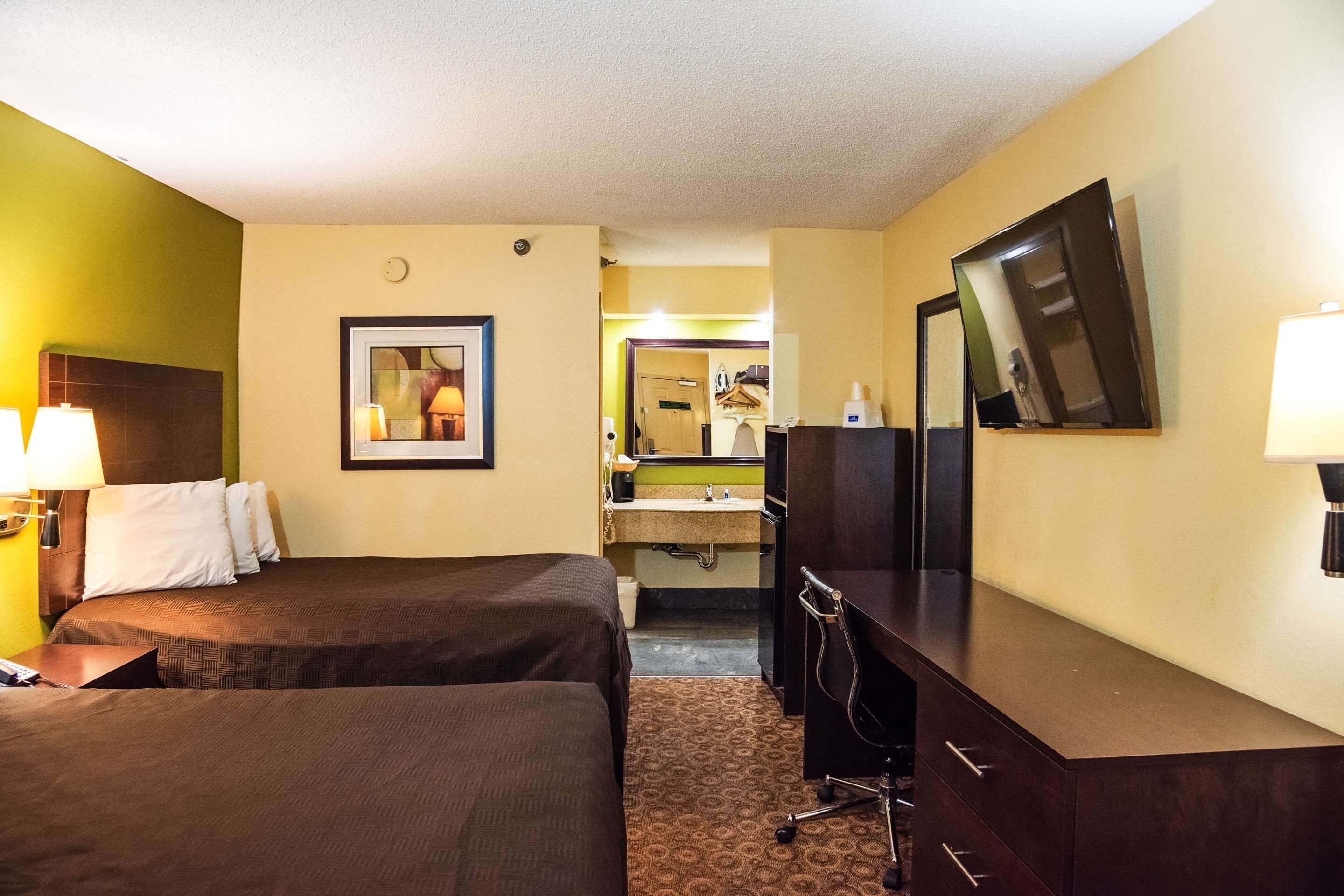 Une chambre d’hôtel avec deux lits, un bureau, un miroir et une salle de bain visible à travers une porte ouverte.