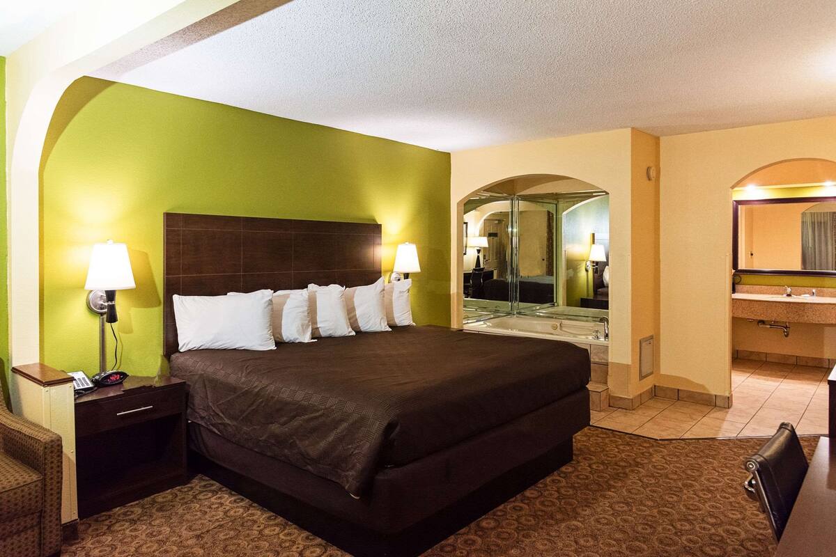 A hotel room with a large bed, two bedside lamps, a nightstand with a phone, a mirror, and a bathroom with a sink and mirror.