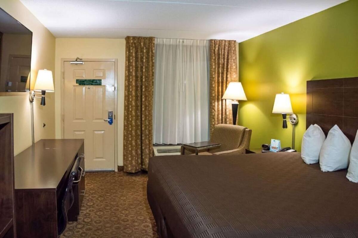 A hotel room with a bed, a desk, a chair, a lamp, and a door.