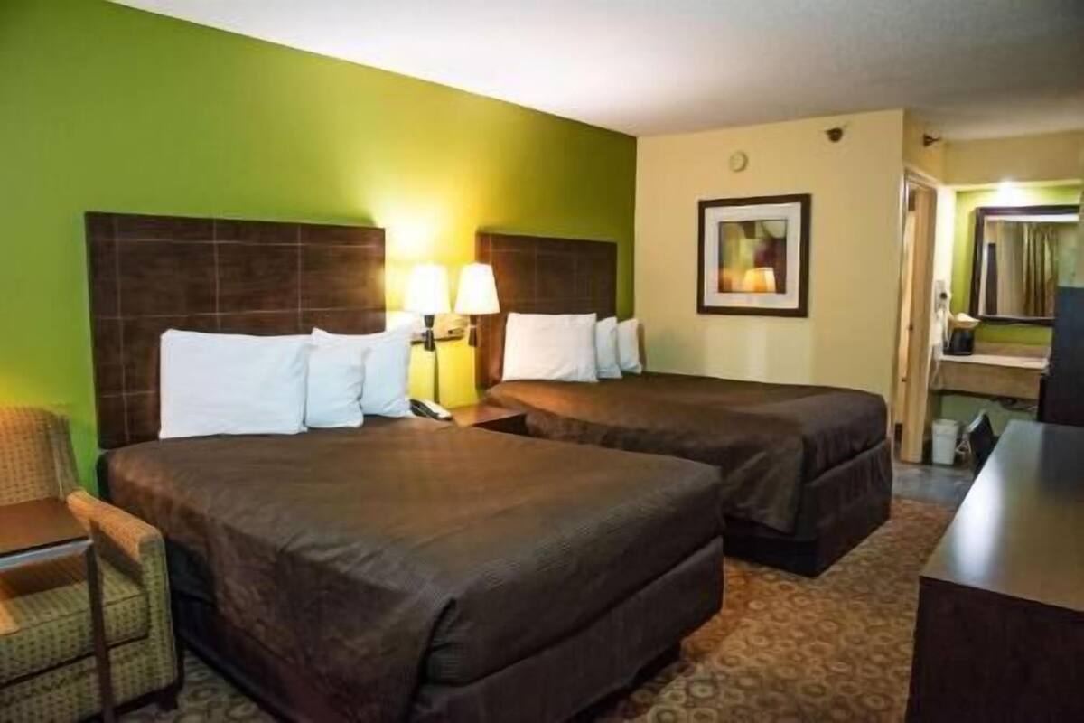 A hotel room with two beds, a desk, a chair, and a lamp.
