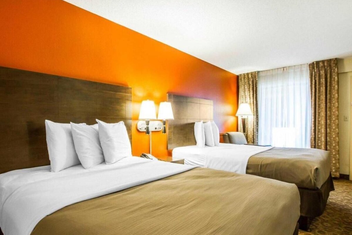 A hotel room with two beds, a wooden headboard, a nightstand, a lamp, and a window with curtains.