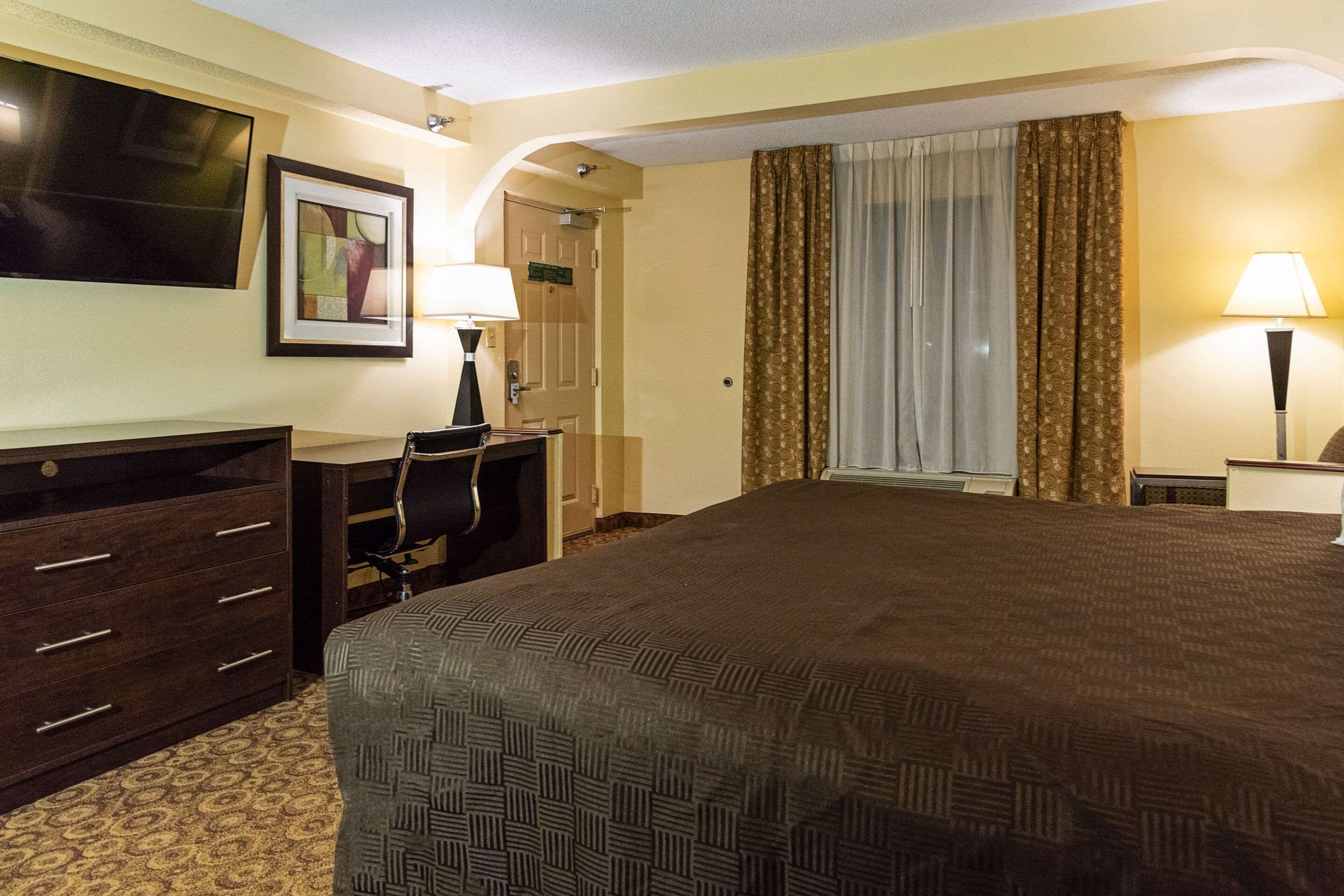 A hotel room with a large bed, a desk with a chair, a TV mounted on the wall, and a window with curtains.