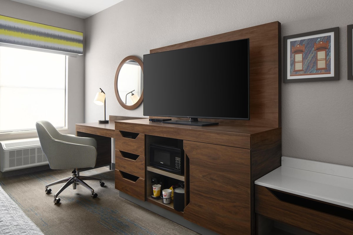 A modern hotel room with a large flat-screen TV mounted on a wooden cabinet, a desk with a chair, a round mirror, and framed artwork on the wall.
