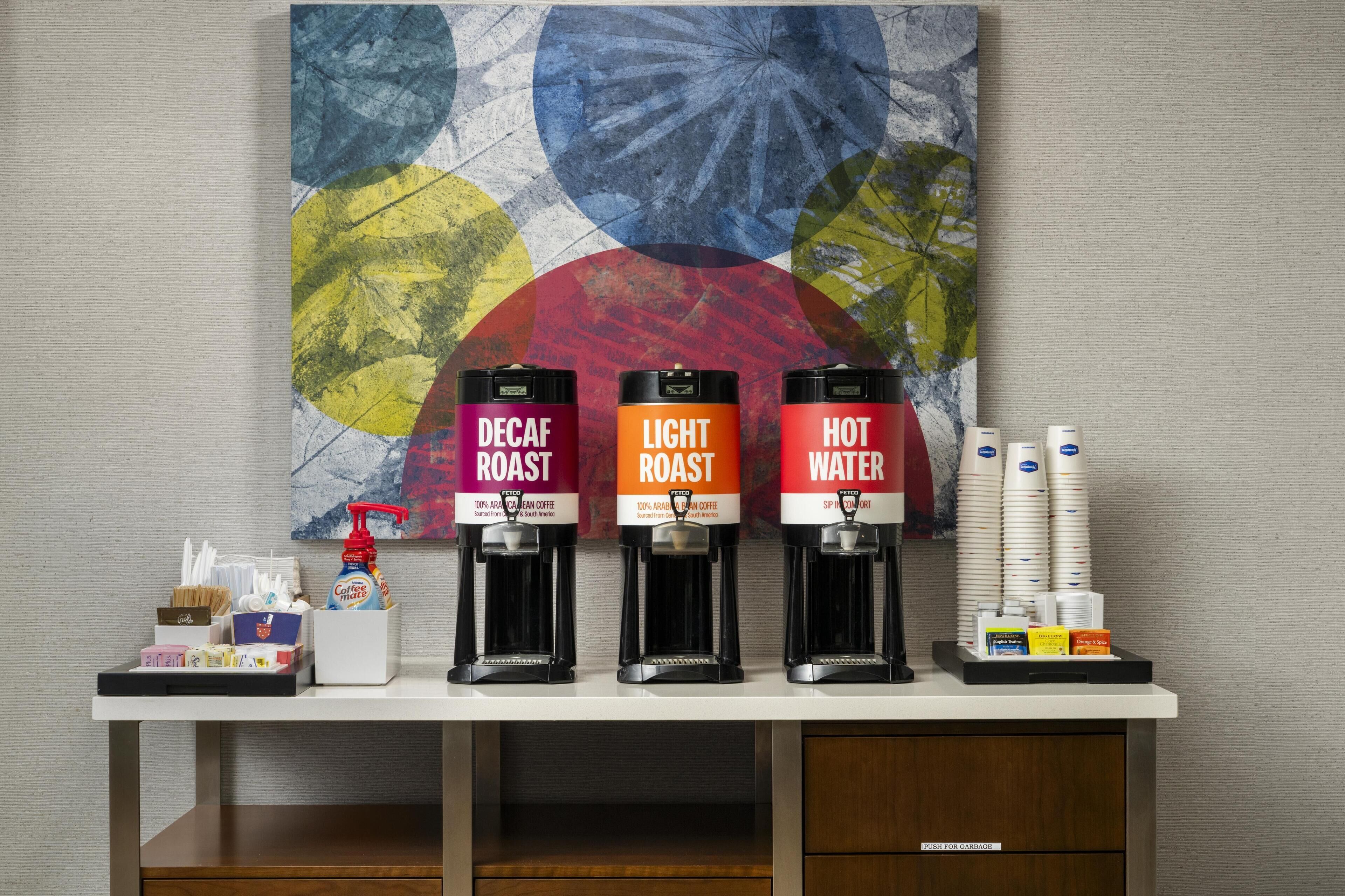 Un coin café équipé de trois distributeurs étiquetés « café décaféiné », « café léger » et « eau chaude ». On y trouve également des piles de gobelets jetables et une boîte de mouchoirs.