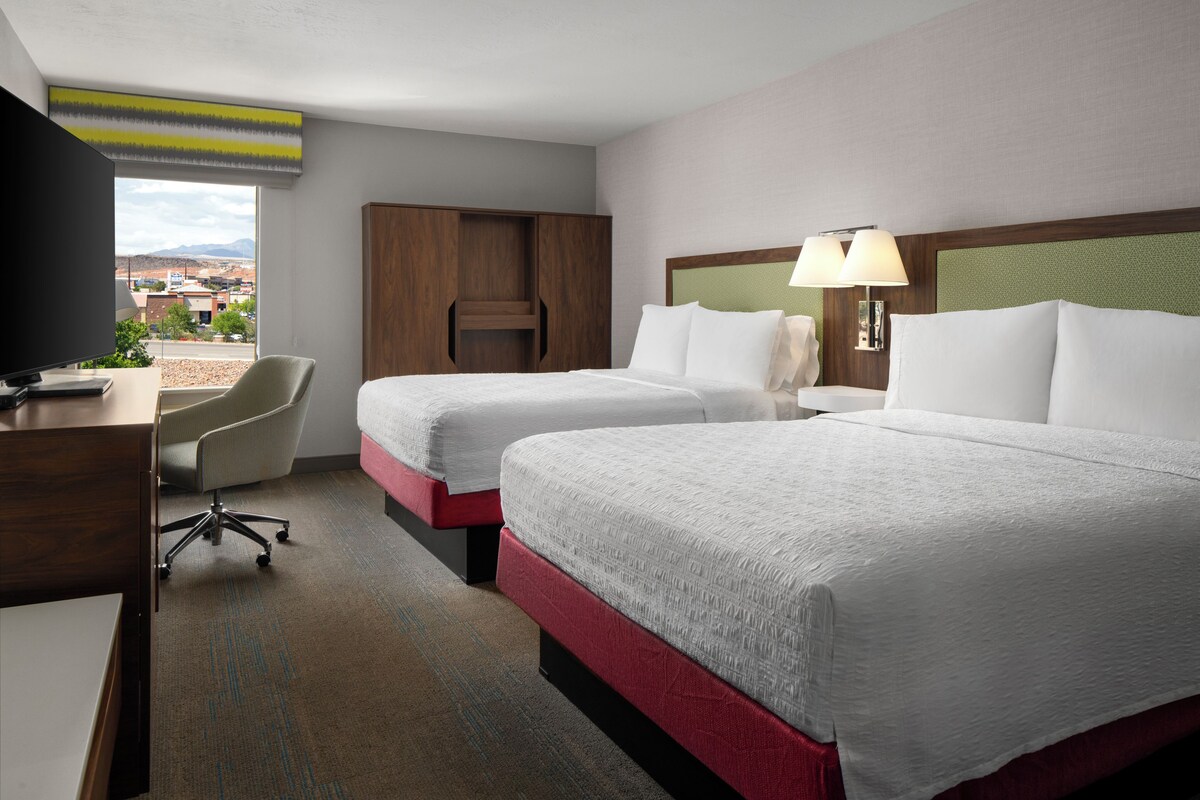 Quarto de hotel com duas camas, uma escrivaninha, uma cadeira, televisão e vista para o exterior.