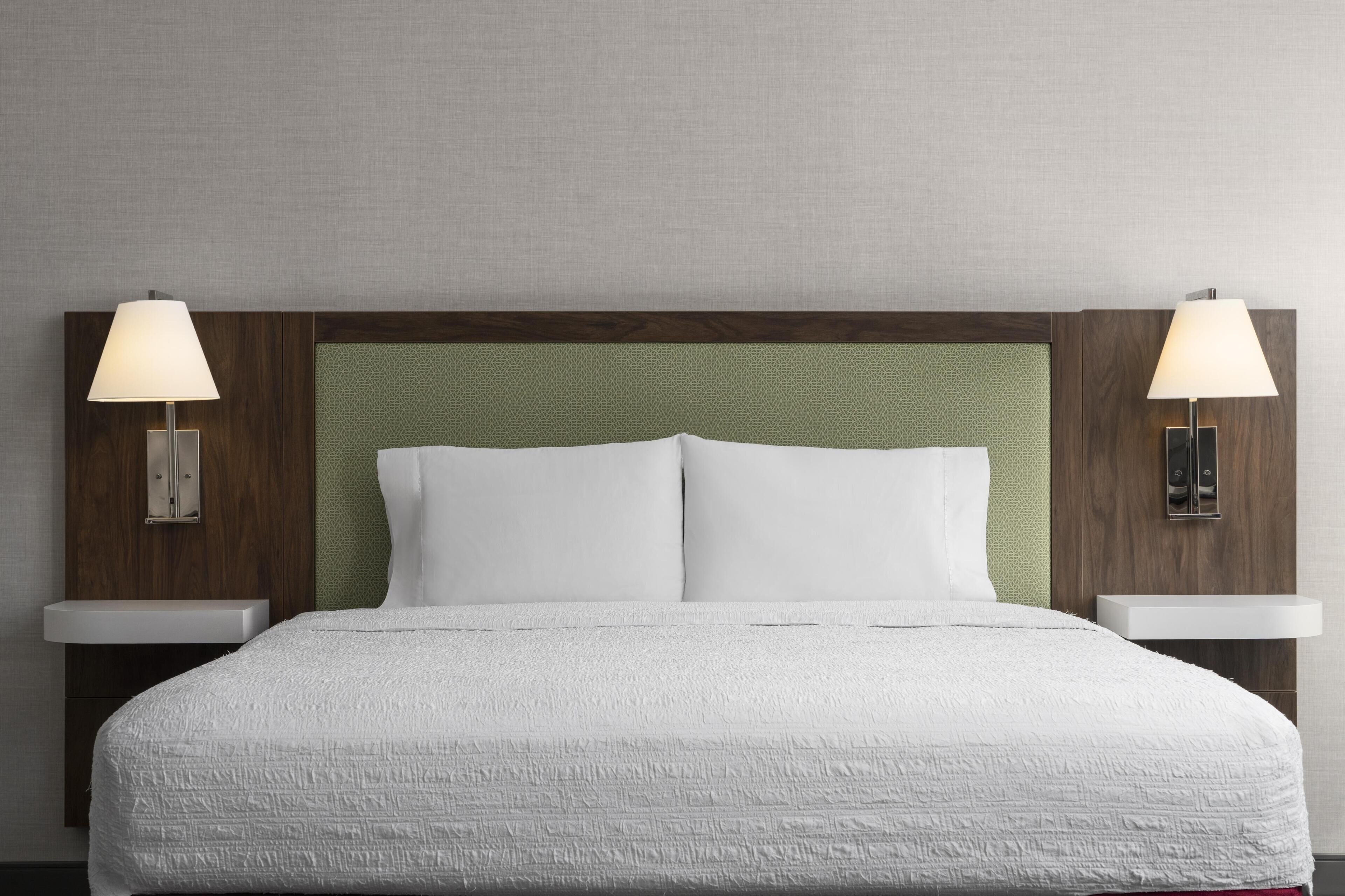 A neatly made bed with a textured white duvet, two bedside lamps, and a wooden headboard with a green panel.