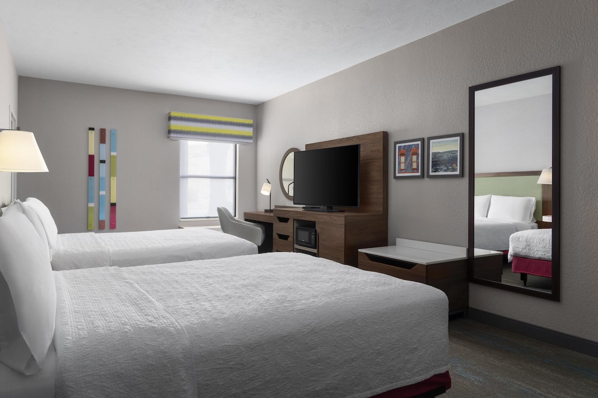 A hotel room with two beds, a TV, a desk, a mirror, and artwork on the wall.