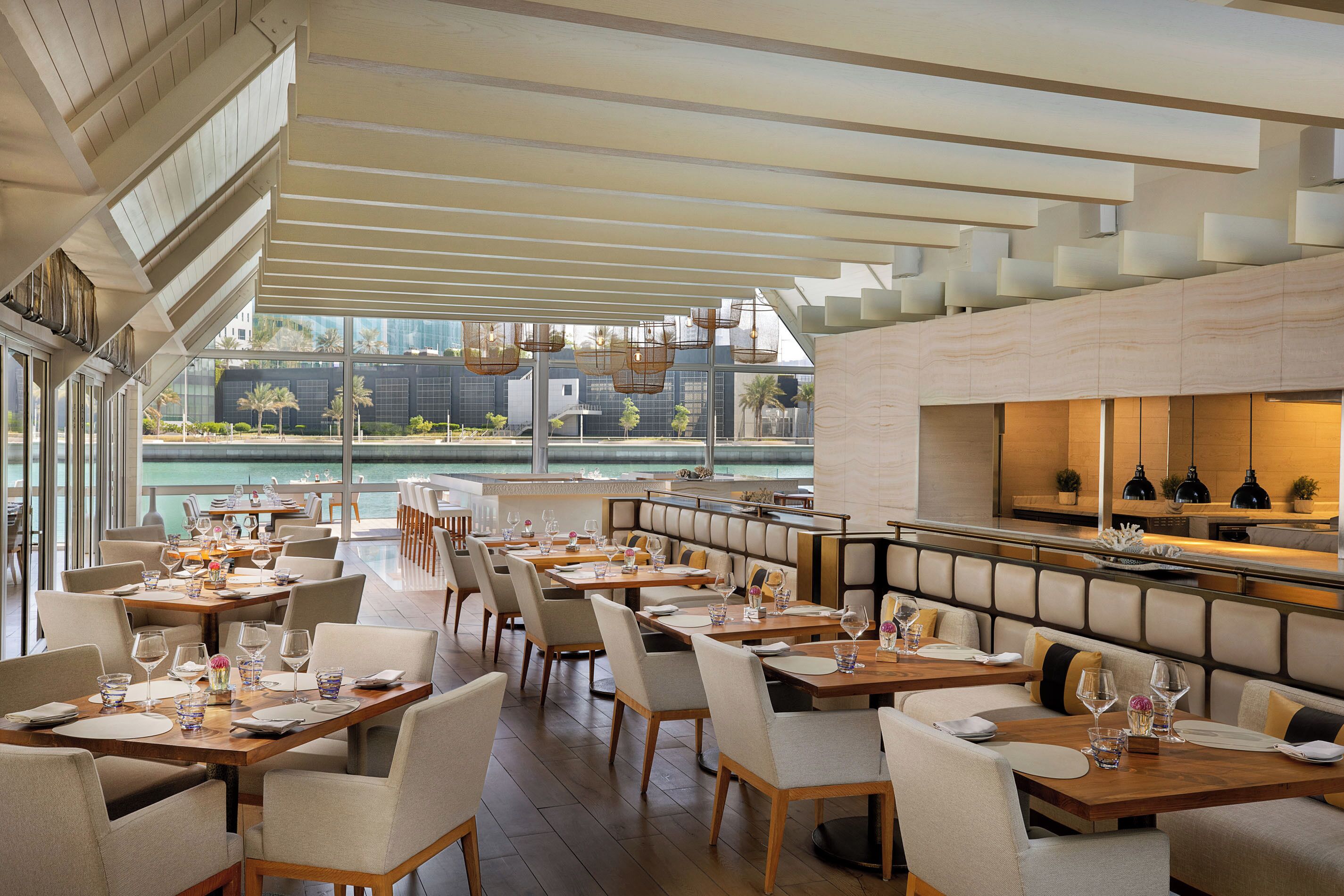 A modern restaurant with high ceilings, large windows, and a view of the water. Tables are set with white tablecloths and chairs.