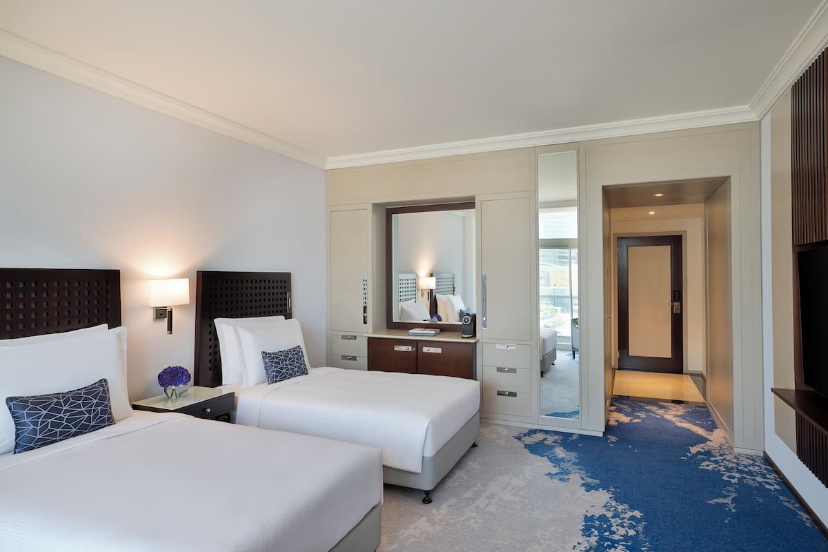 A hotel room with two beds, a large mirror, a desk with a lamp, and a view of the city through the window.