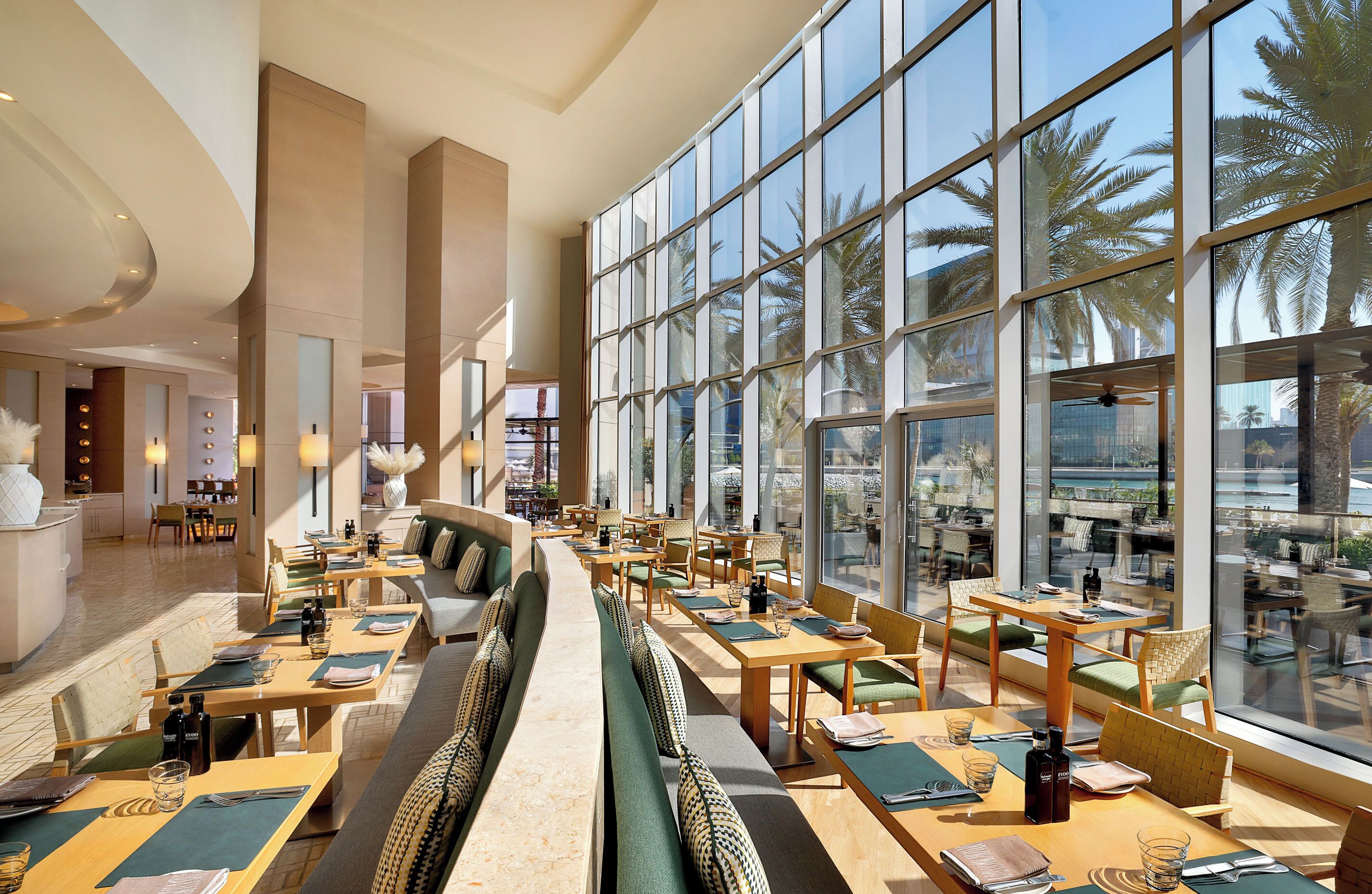 A spacious indoor dining area with high ceilings, large windows, and a view of palm trees.