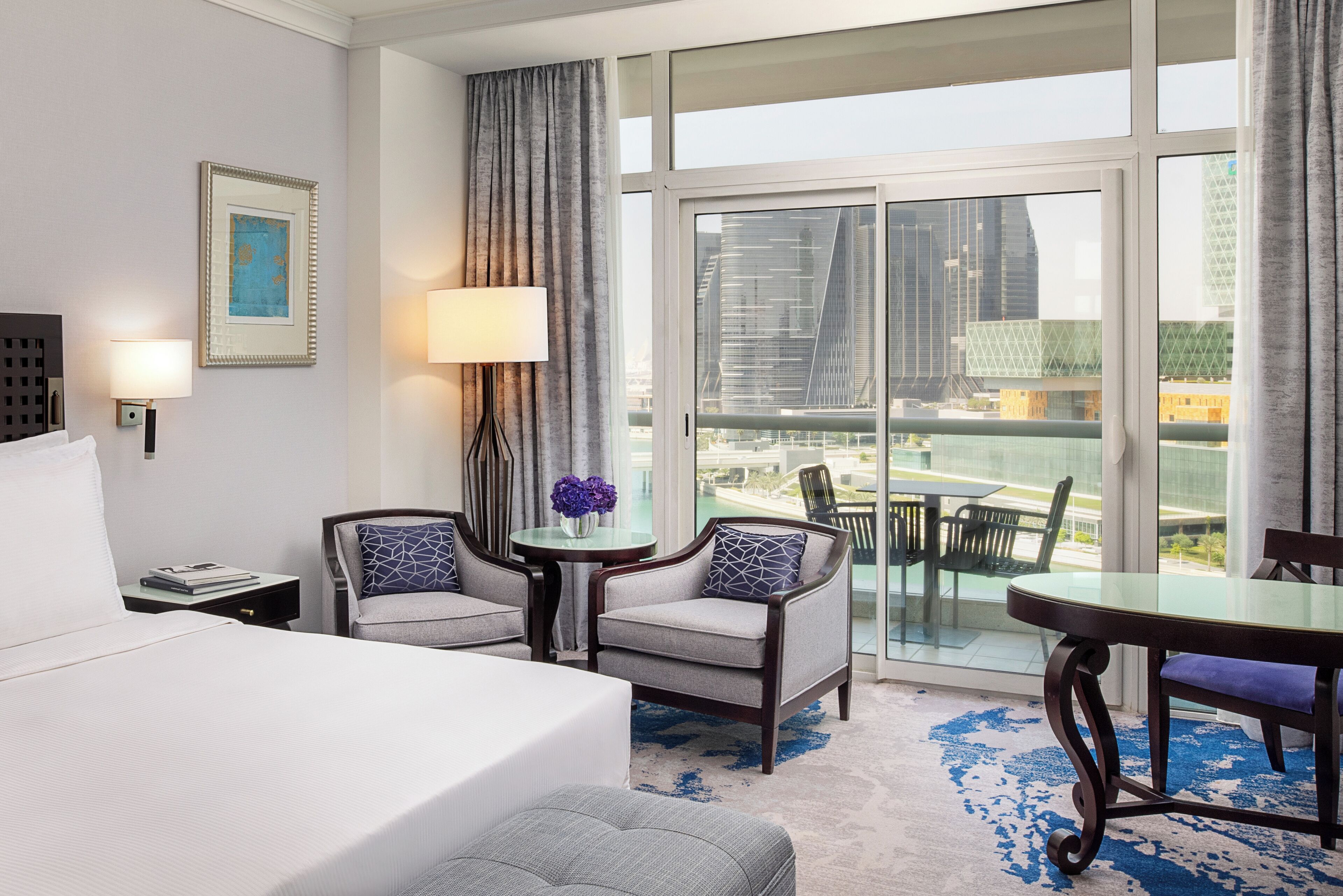 A hotel room with a bed, two armchairs, a small table with a vase of flowers, a dining table, and a large window with a city view.