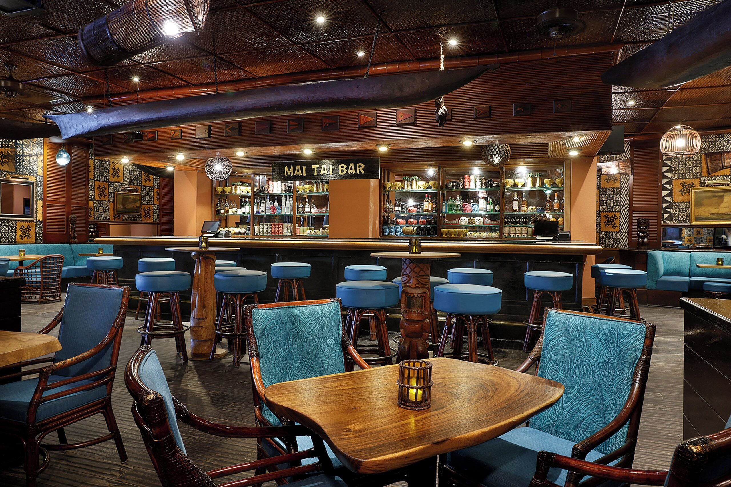 Eine Bar mit Holzsitzmöbeln, blau gepolsterten Sitzflächen und einer Auswahl an Flaschen und Gläsern auf Regalen.