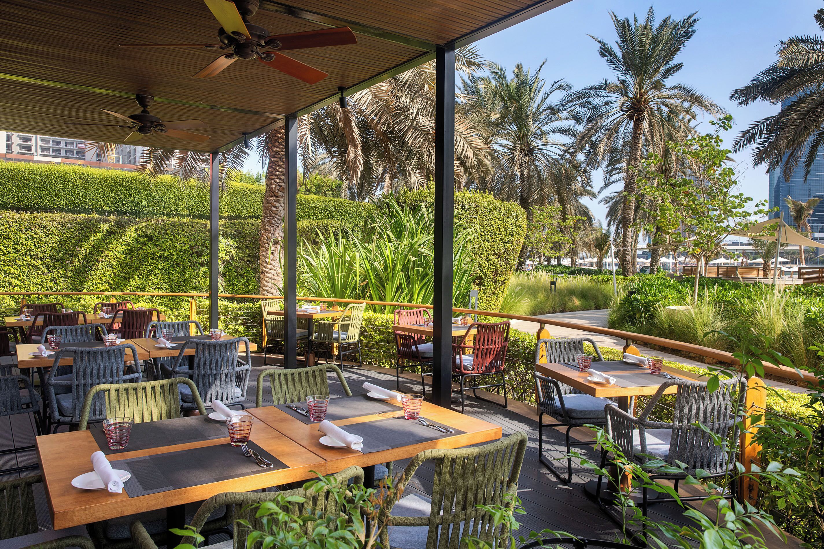 An outdoor restaurant with wooden tables and chairs, surrounded by lush greenery and palm trees.