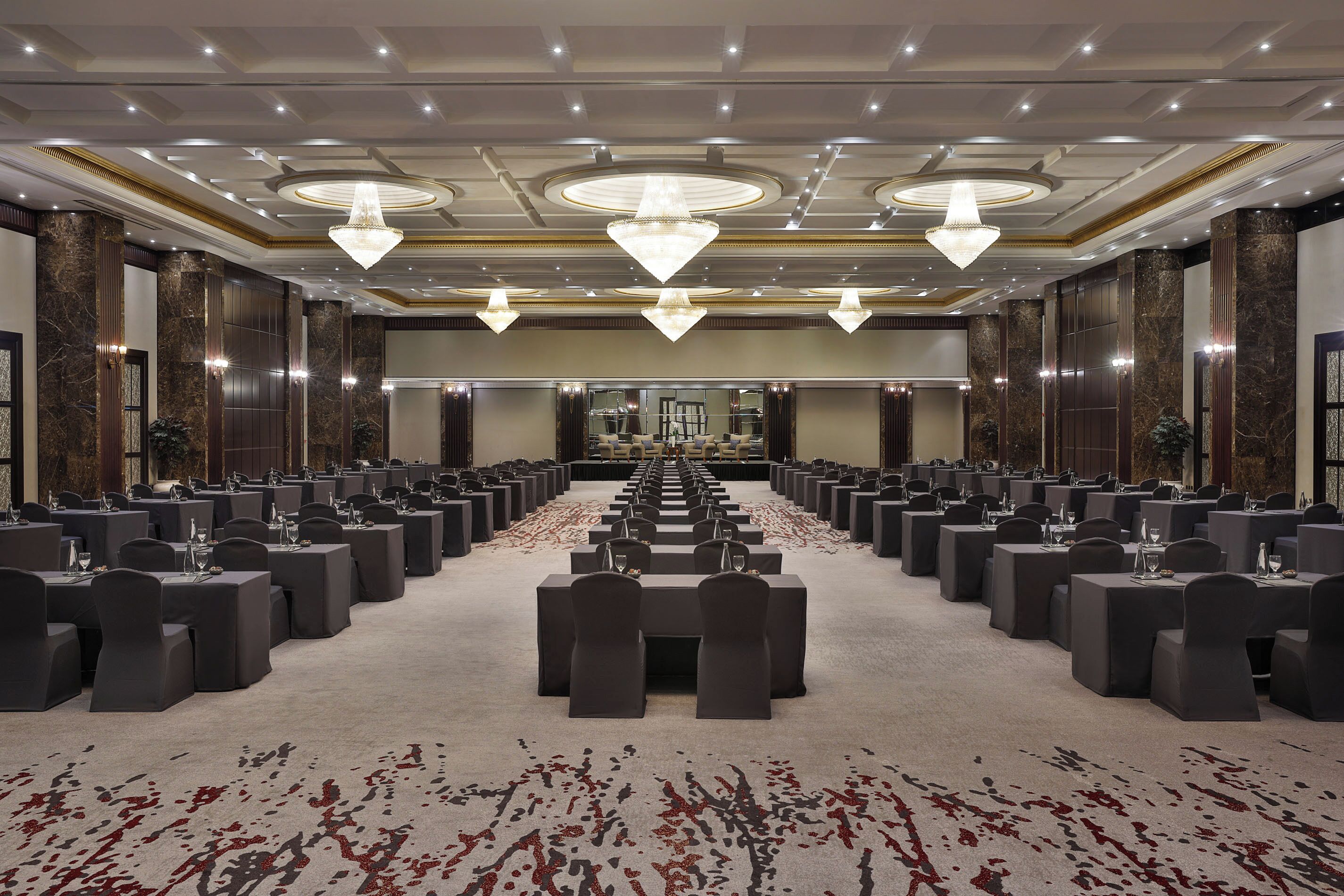 A spacious conference room with rows of tables and chairs, large chandeliers, and a carpeted floor.