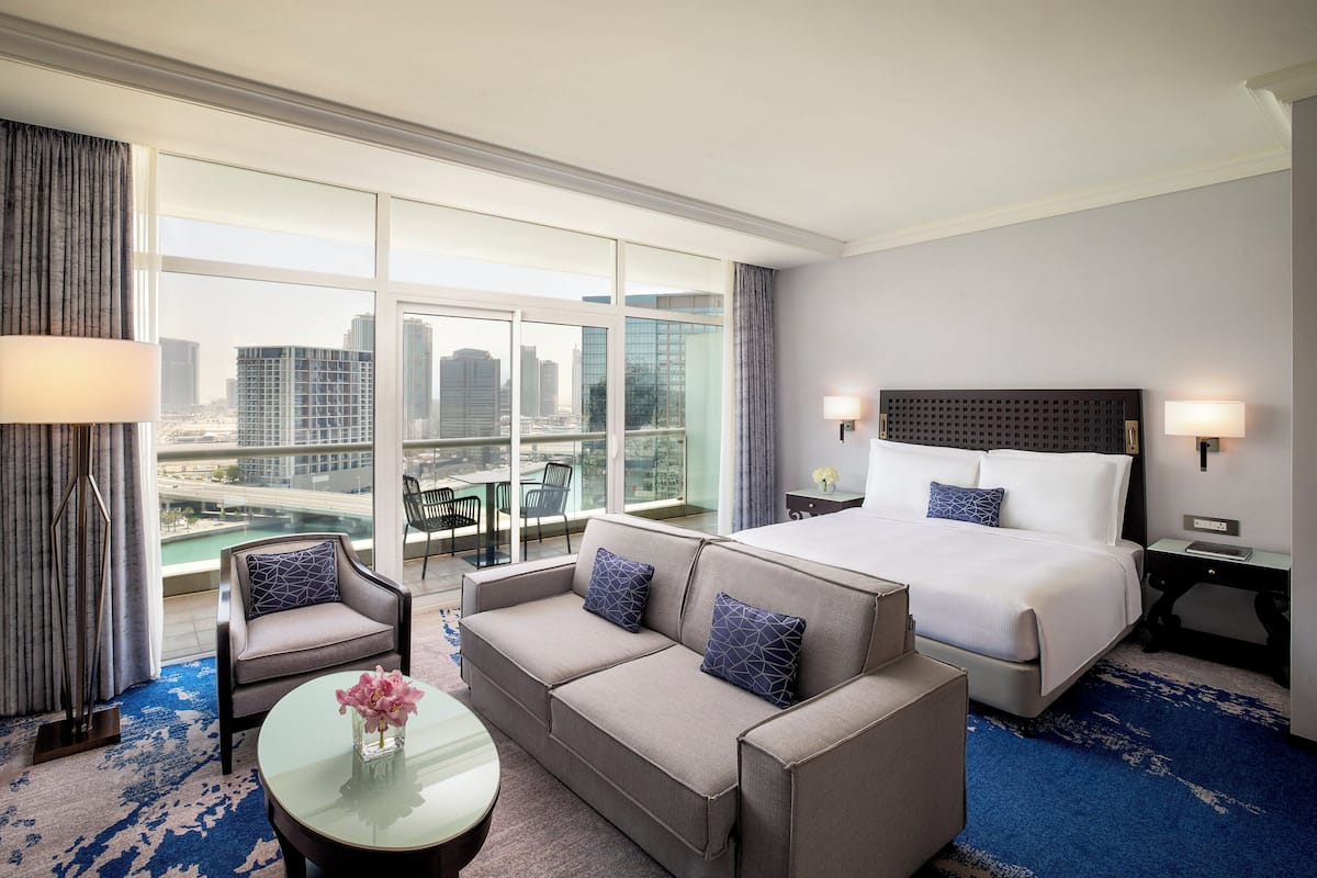 A hotel room with a large bed, a sofa, a small table with a vase, and a view of the city through large windows.