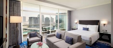 A hotel room with a large bed, a sofa, a small table with a vase, and a view of the city through large windows.