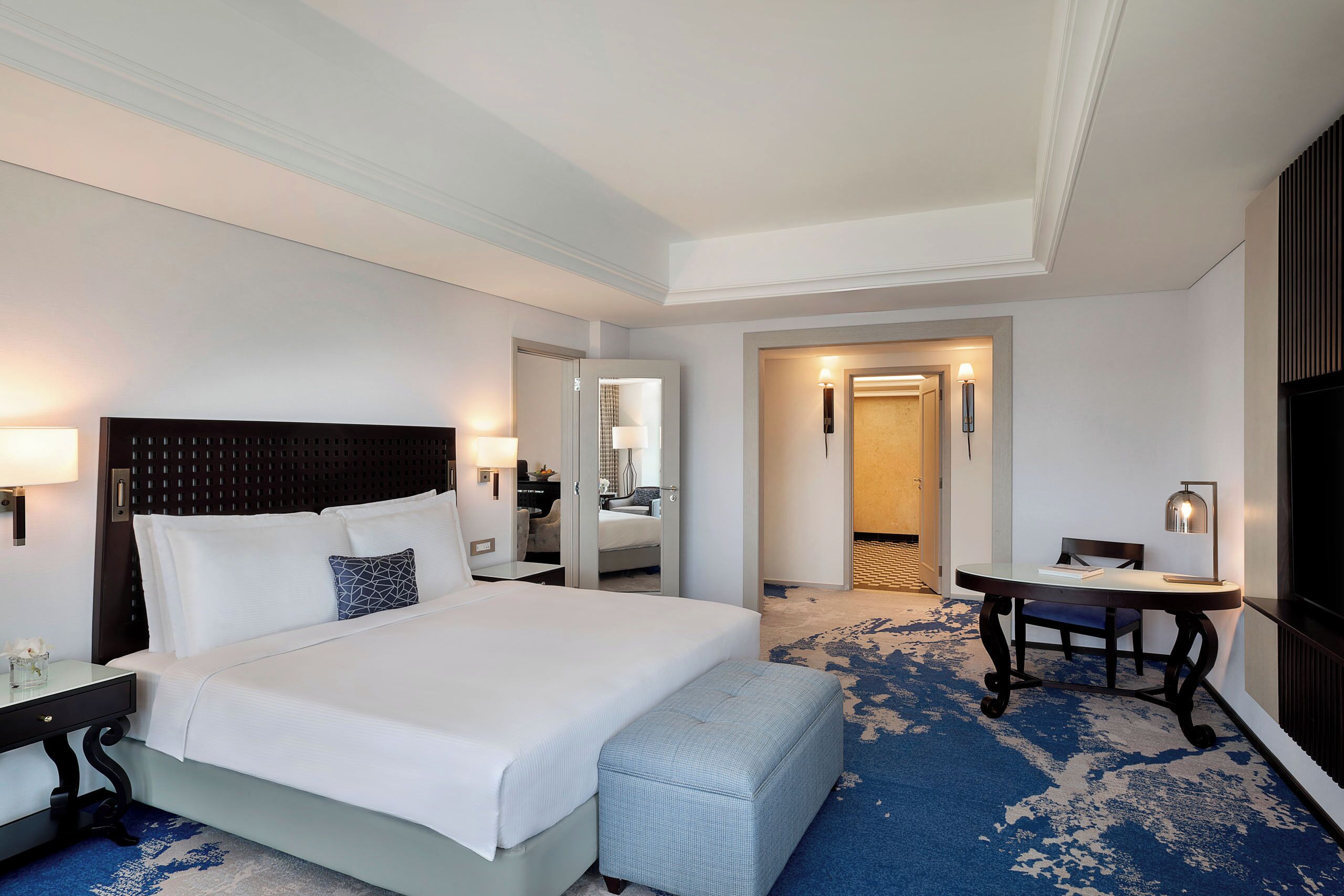 A hotel room with a large bed, a desk, a chair, and a small table with a lamp.
