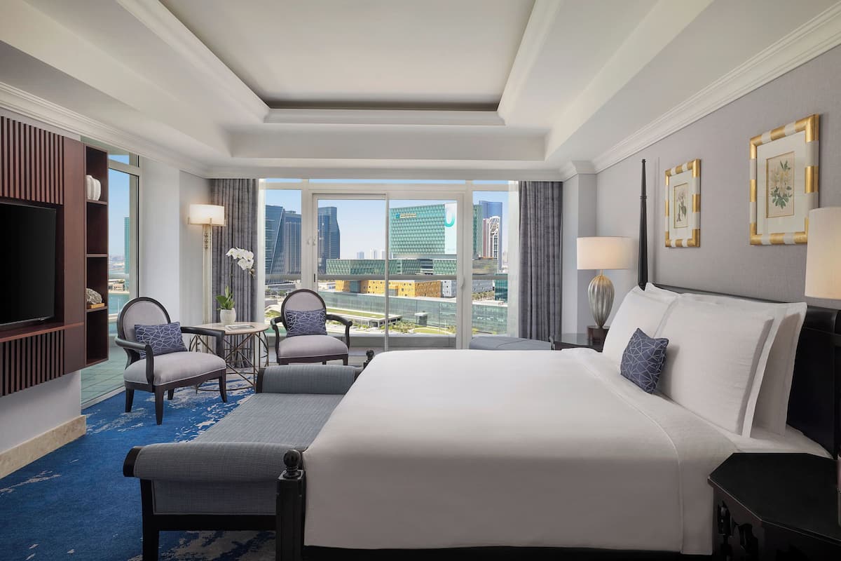 A hotel room with a large bed, two armchairs, a small table, and a TV. The room has large windows with a city view.