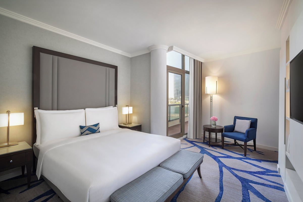 A hotel room with a large bed, bedside tables with lamps, a small round table with a chair, and a TV on the wall.